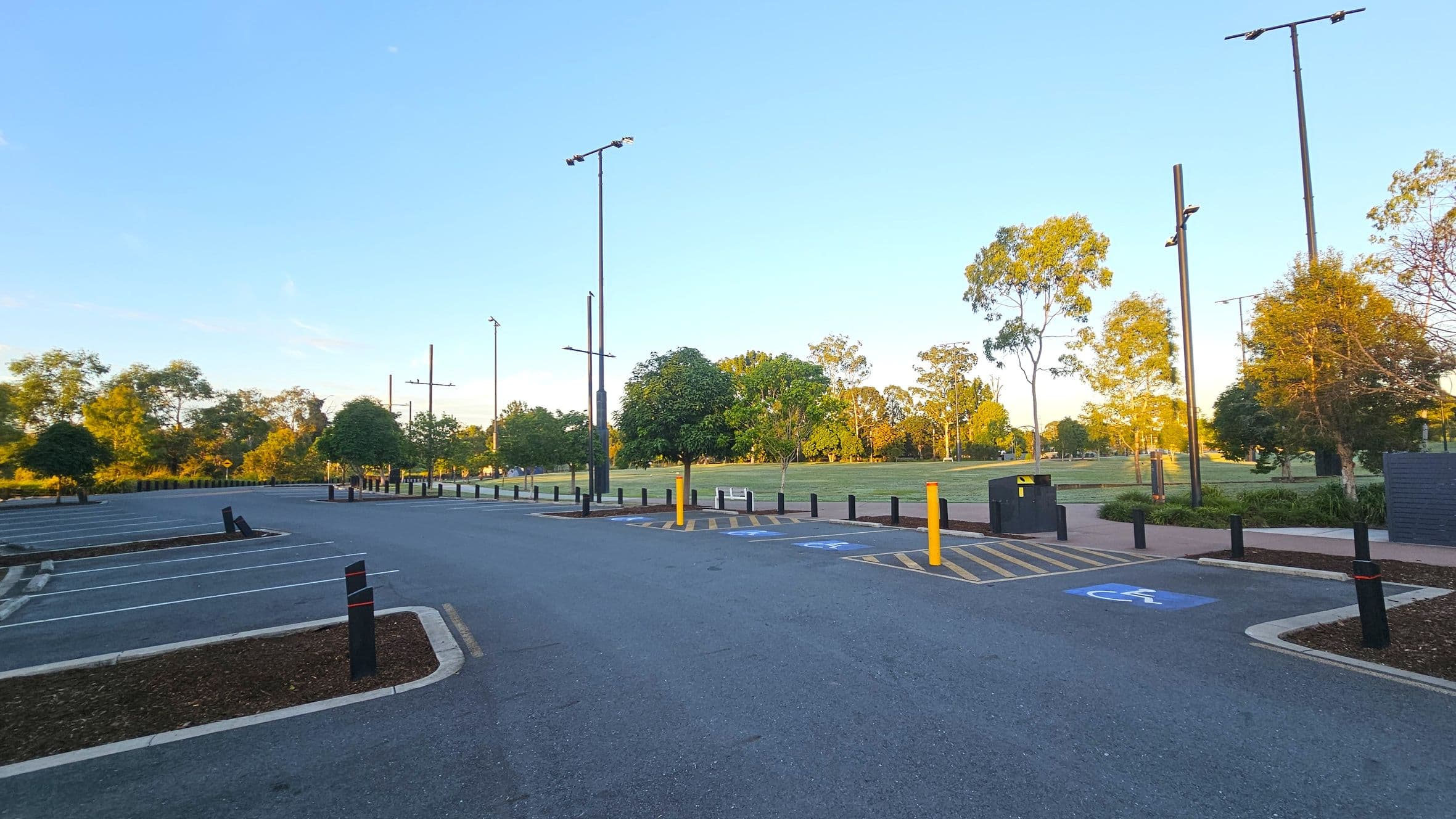 A car park with marked spaces, including a designated disabled parking spot on the right. Tall light poles line the area. In the background, a grassy field is bordered by trees. The ground is paved, and there are small landscaped sections with mulch and plants.
