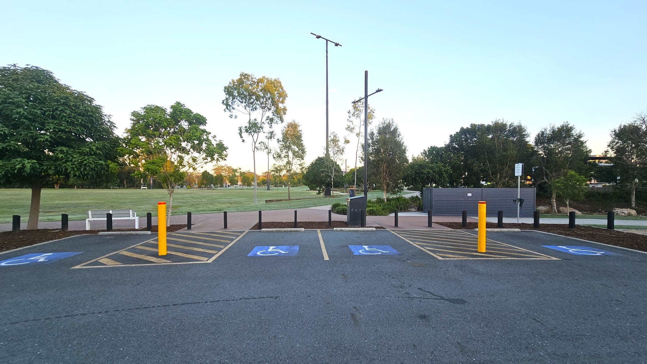 A parking area with two marked disabled parking spaces on an asphalt surface. Yellow lines and a yellow bollard separate the spaces. In the background, a grassy park with scattered trees and a white bench is visible. Tall streetlights and a grey utility box are also present. A paved path runs parallel to the parking area, bordered by black bollards.