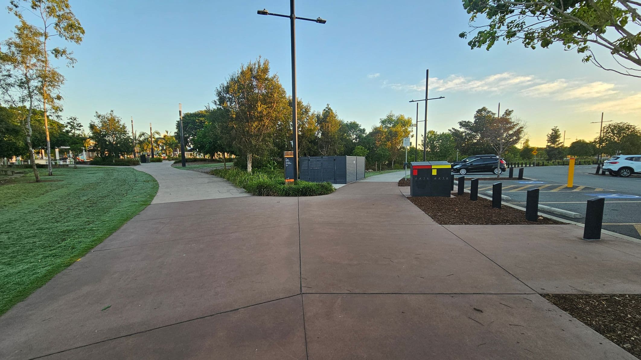 A paved path splits into two directions, with grass and trees lining both sides. On the left, a grassy area with scattered trees and a few benches. On the right, a car park with several vehicles and a colourful utility box. Tall streetlights are positioned along the paths. In the background, more trees and a clear sky. The ground is a mix of concrete and grass, with some mulch around the utility box.