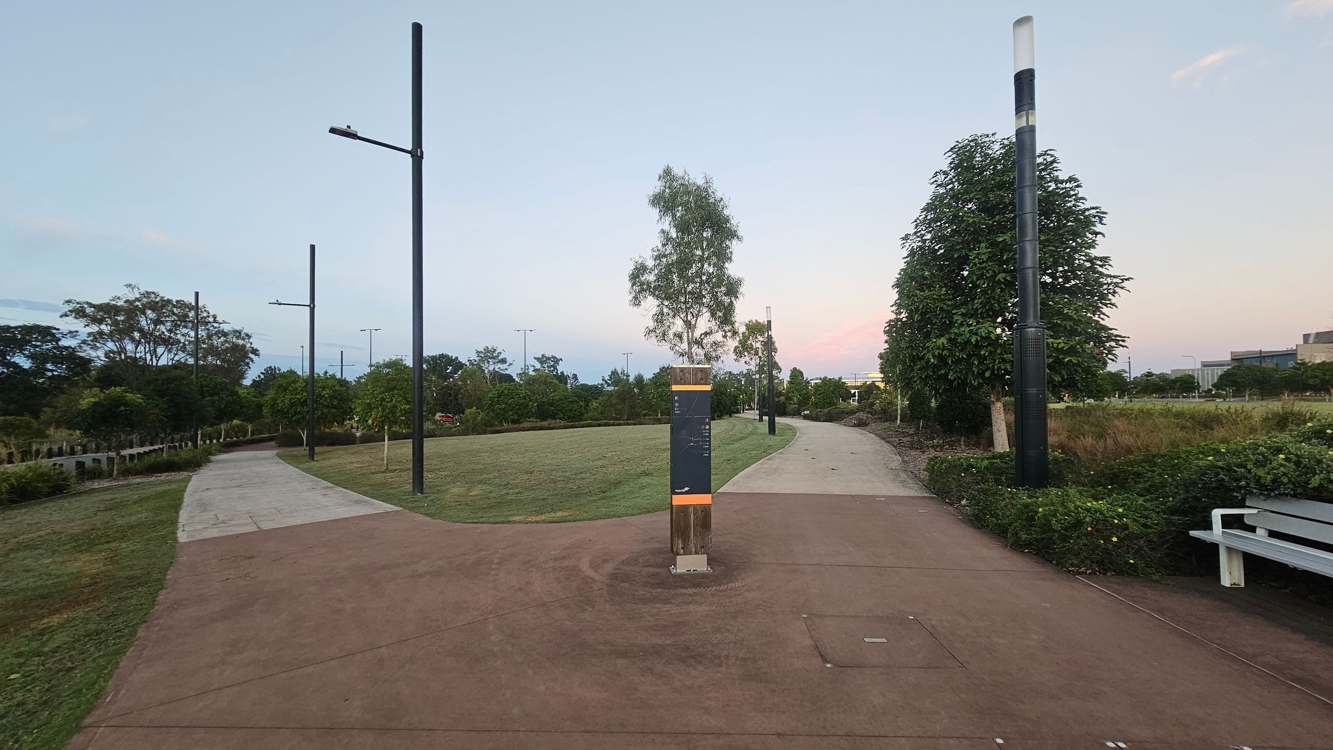 A park scene with a paved path splitting into two directions. In the centre, a signpost stands on the path. On the left, the path curves with several tall streetlights and trees lining it. On the right, the path is bordered by bushes and a large tree, with a white bench nearby. The background features more trees and a building in the distance. The ground is covered with grass, and the sky is clear.