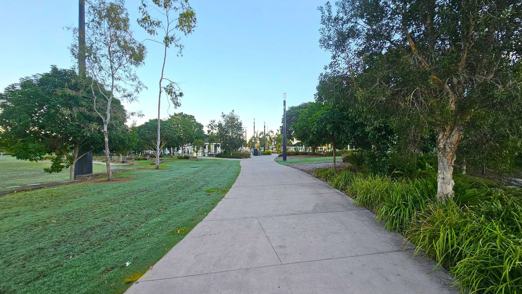 A concrete path runs through the centre, flanked by green grass and trees on both sides. On the left, there are several trees and a grassy area. On the right, more trees and bushes line the path. In the background, there are more trees and some structures, possibly part of a park or garden area. The sky is clear and blue.