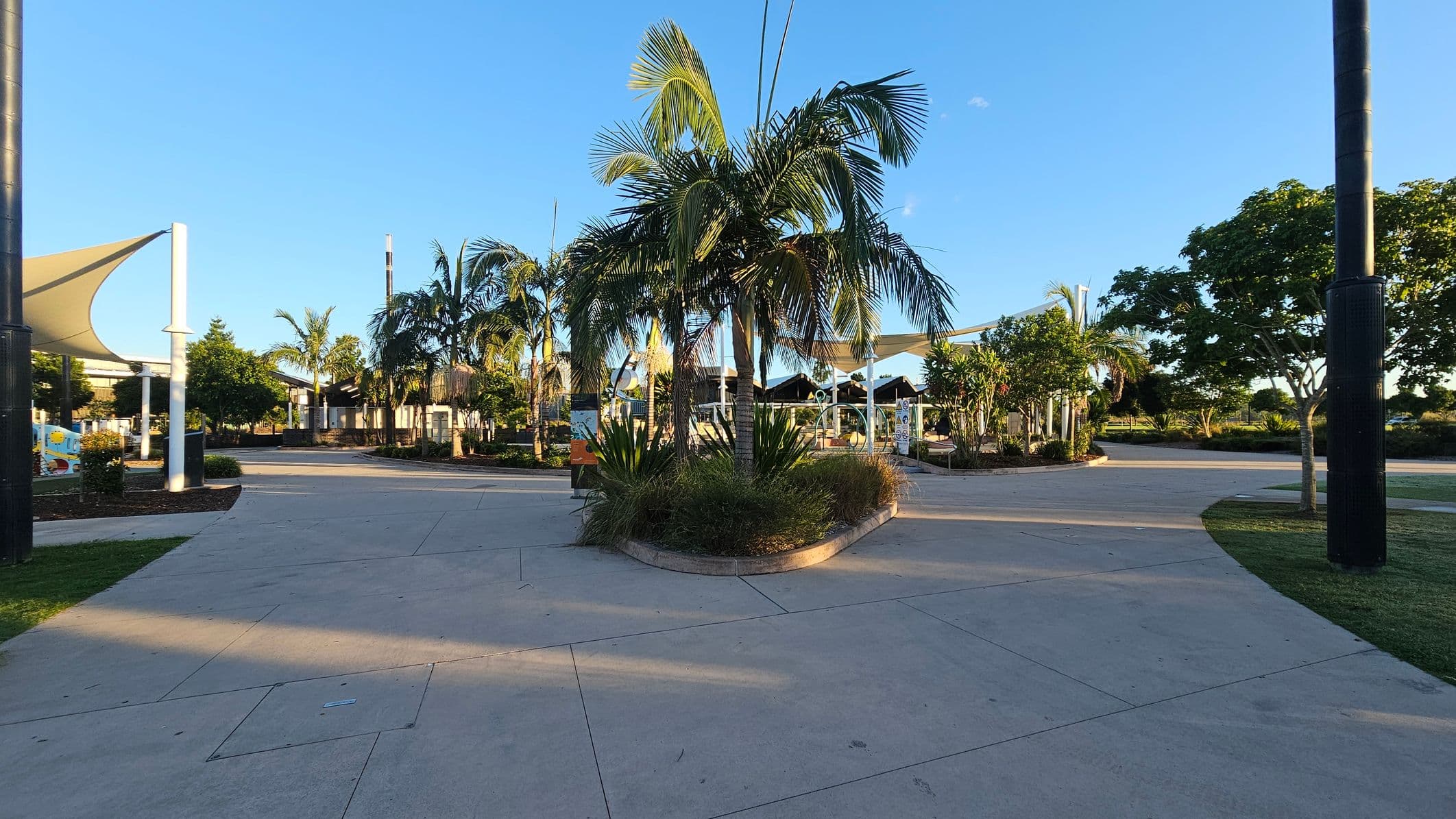 A paved area with a central island of palm trees and shrubs. On the left, there are white poles supporting triangular shade sails. To the right, more trees and greenery are visible, with a large black pole in the foreground. The background features additional trees and a clear blue sky. The ground is a light-coloured concrete with visible seams.