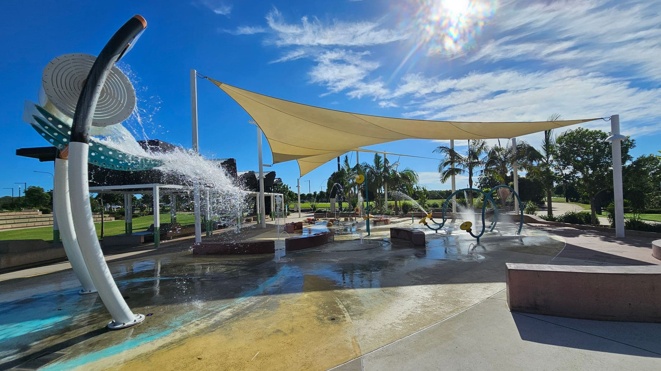 A water play area with a large, curved water feature on the left, spraying water onto the ground. In the centre, there are various water play structures, including circular frames and small fountains. A large triangular shade sail is overhead, providing cover. Palm trees and greenery are visible in the background. The ground is wet and colourful, with a mix of blue and yellow tones. Benches are scattered around the area.