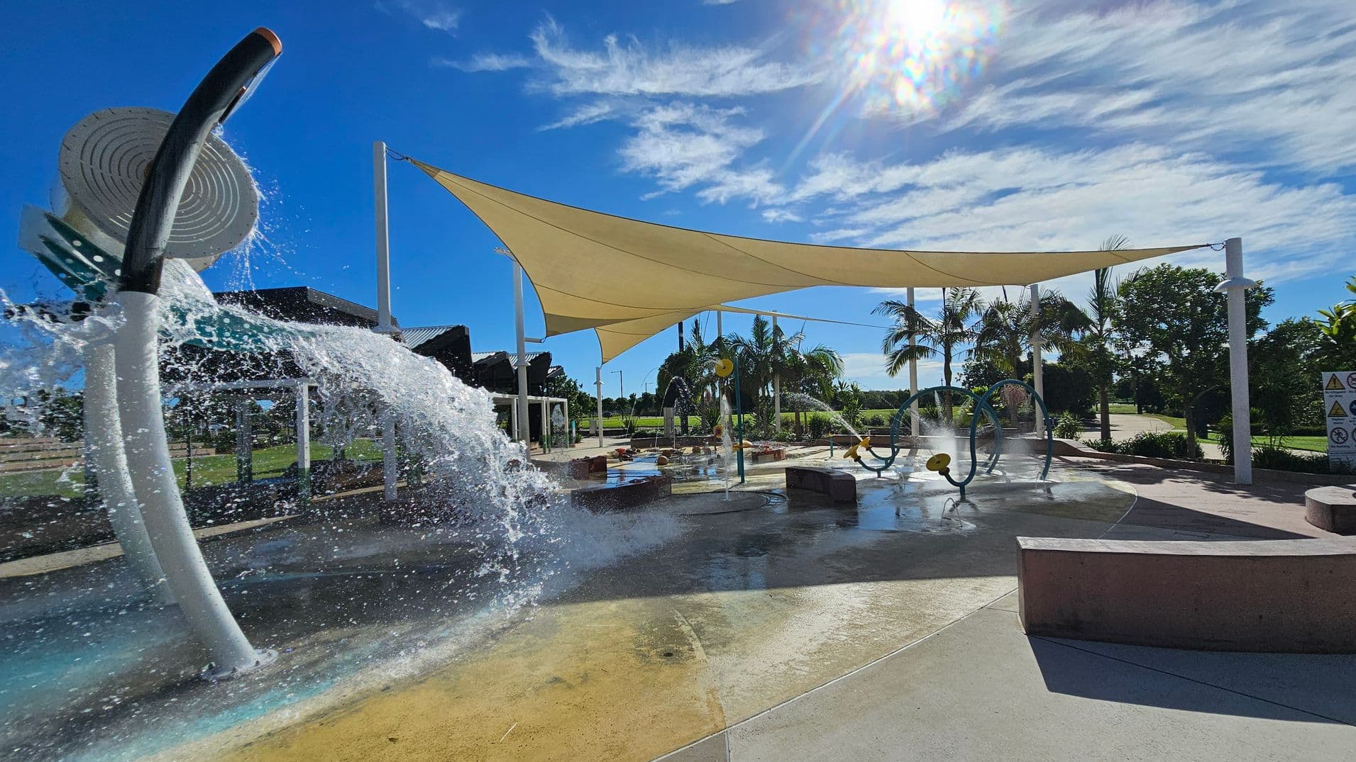 A water play area with a large, curved water feature on the left, spraying water into the air. In the centre, there are various water play structures, including circular hoops and small fountains. A large beige shade sail is stretched overhead, supported by white poles. Palm trees and greenery are visible in the background. The ground is wet and made of smooth concrete, with some colourful sections. The sun is shining brightly, casting shadows across the area.