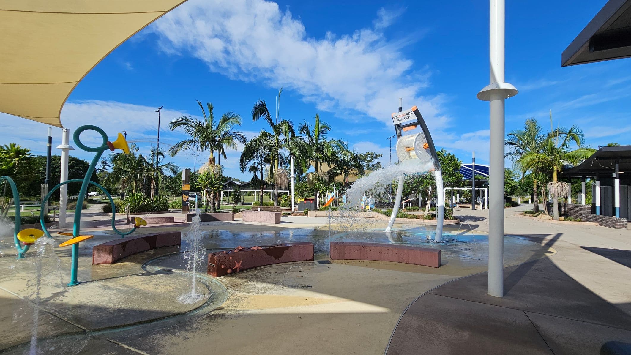 A water play area with various interactive features, including a large bucket pouring water and colourful pipes with yellow accents. Palm trees and greenery are in the background, providing a tropical feel. The ground is wet, indicating recent water activity. A large shade structure is visible on the left, and a building with a dark roof is on the right. The area is open and spacious, with clear pathways and seating areas.