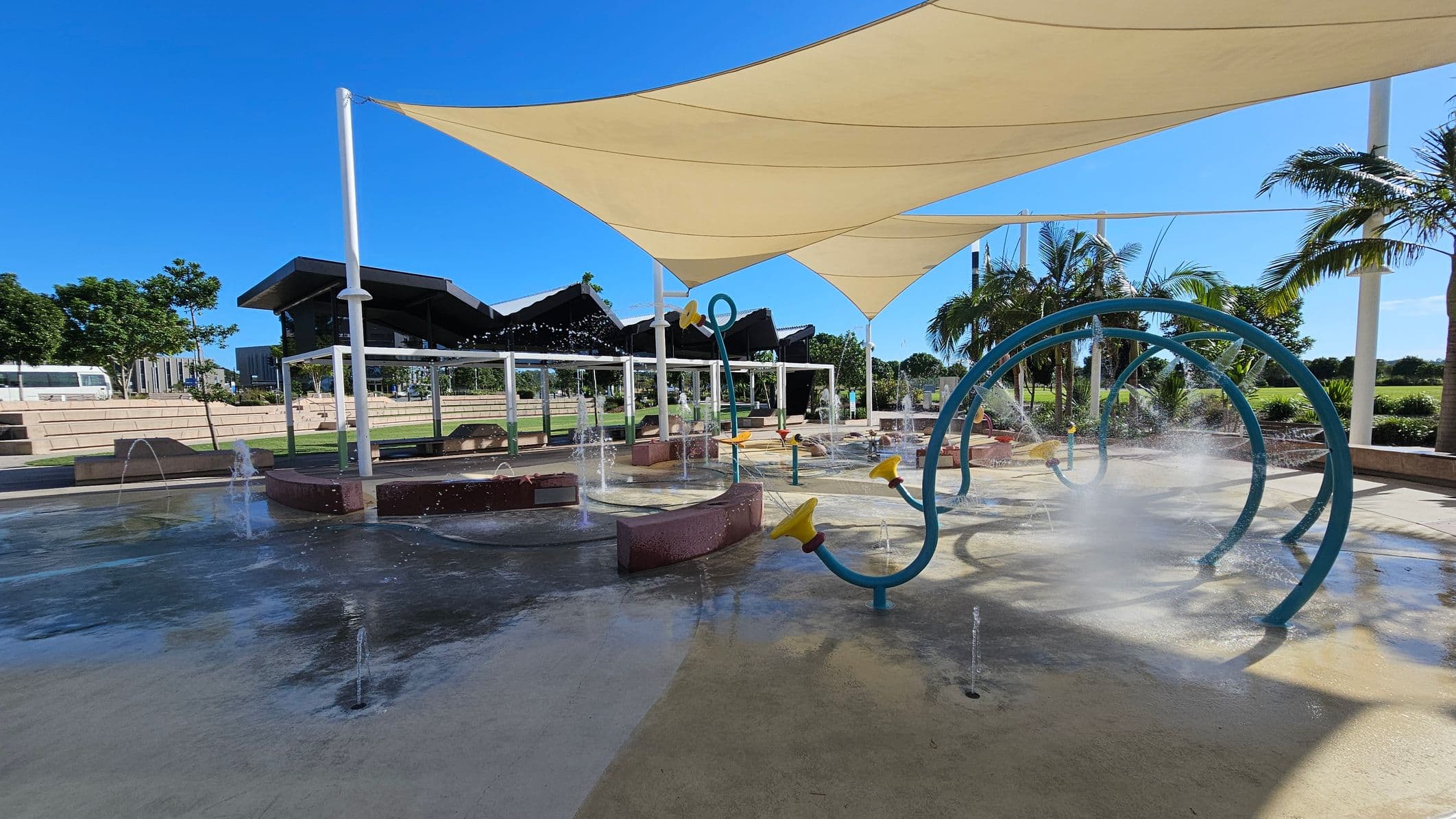 A water play area with various interactive features, including blue circular structures spraying water. Yellow nozzles are attached to some of the structures. The ground is wet with water jets emerging from it. Overhead, large triangular shade sails provide cover. In the background, there are palm trees and a grassy area with a few buildings visible. The sky is clear and blue.