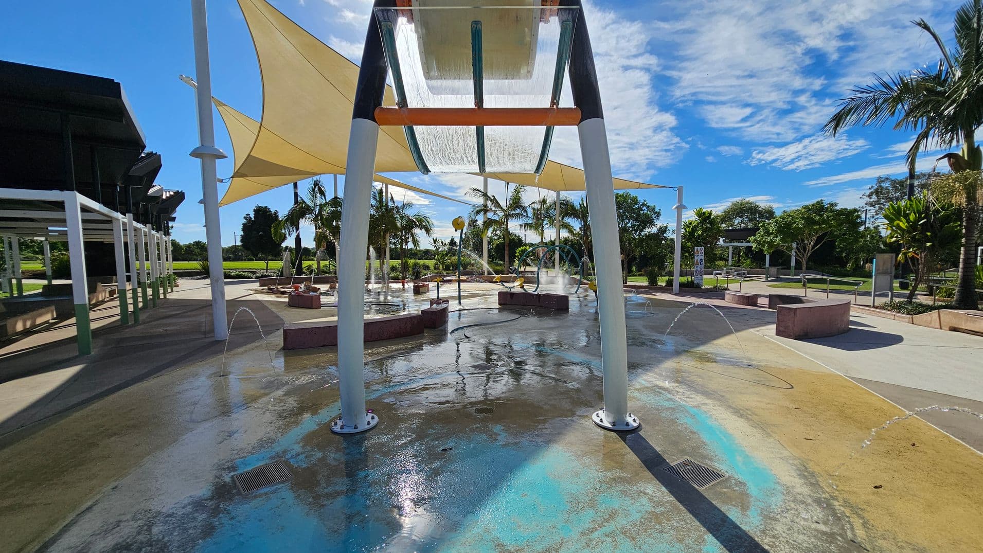 A water play area with a large overhead structure in the centre, featuring a transparent panel with water cascading down. The ground is wet with blue and beige sections. On the left, there are shaded seating areas with white frames. In the background, palm trees and greenery are visible, along with additional play equipment. The sky is partly cloudy, and the area is well-lit by sunlight.