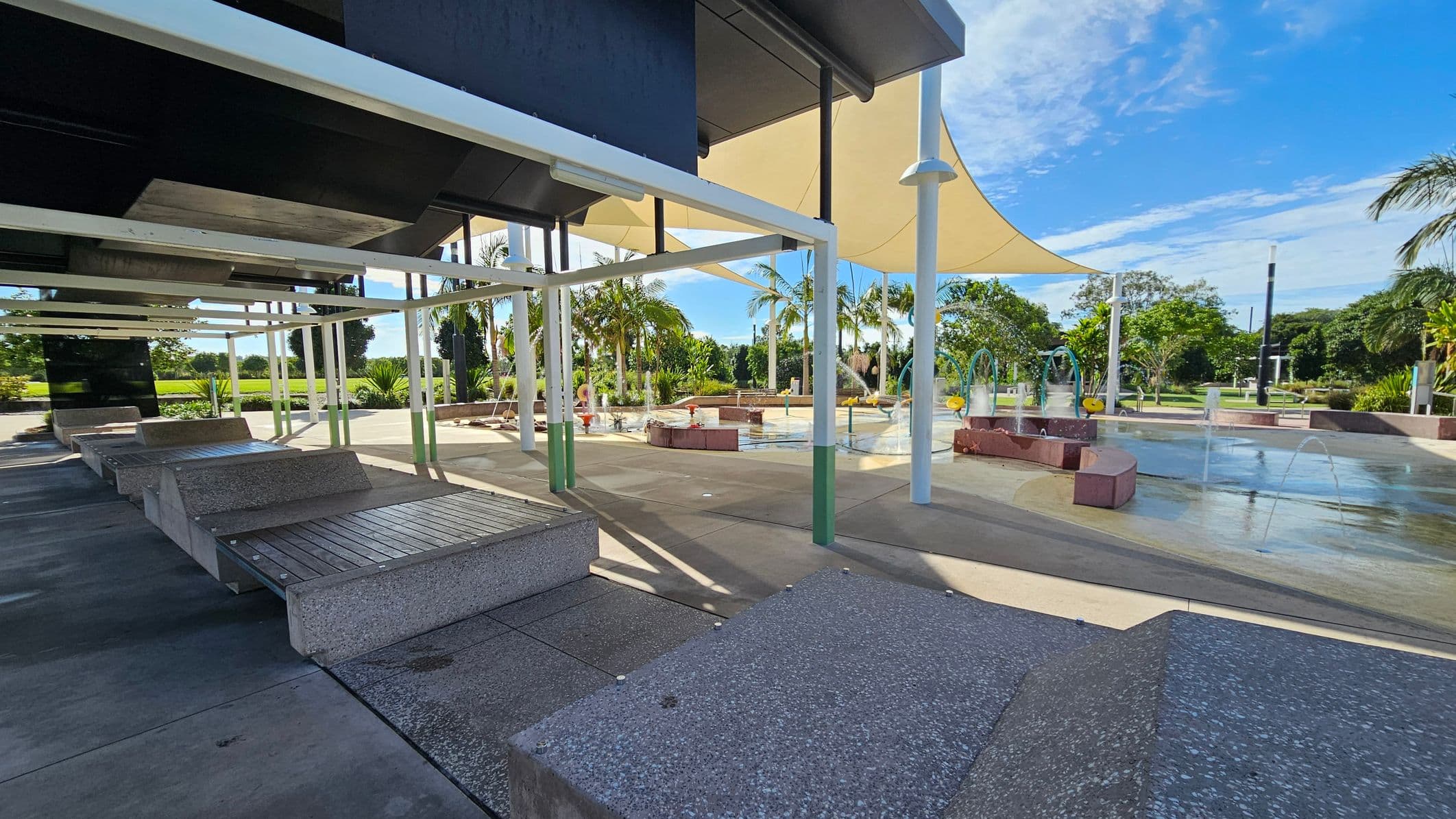 On the left, there are several concrete benches with wooden slats under a shaded structure with white beams. The middle features a paved area with a water play zone, including fountains and splash pads. On the right, there are palm trees and other greenery. The background shows more trees and a clear blue sky. The ground is a mix of concrete and wet surfaces from the water features.