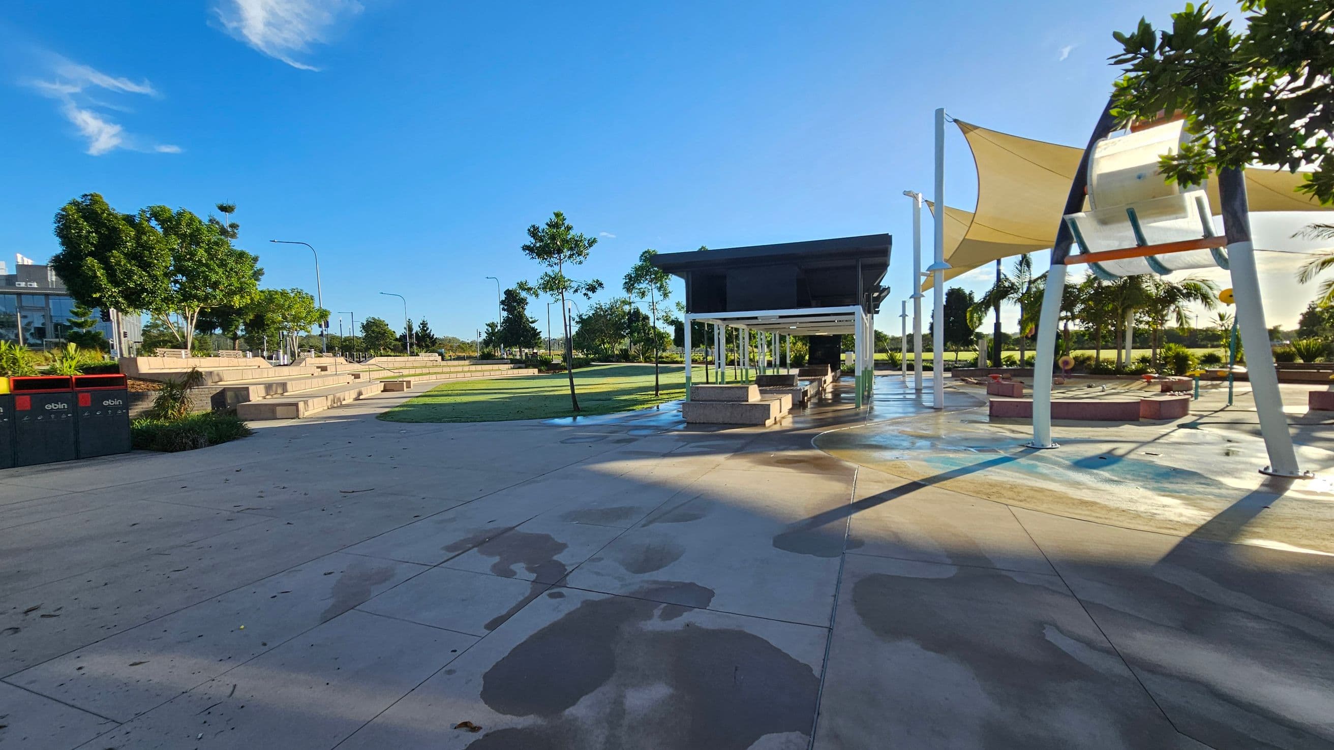A park with a paved walkway in the foreground, featuring scattered wet patches. On the left, there are two black and red bins and tiered seating surrounded by trees and streetlights. In the centre, a white pergola structure with benches underneath. On the right, a large shade sail supported by metal poles, with more trees and greenery in the background. The area is well-lit with clear shadows cast on the ground.