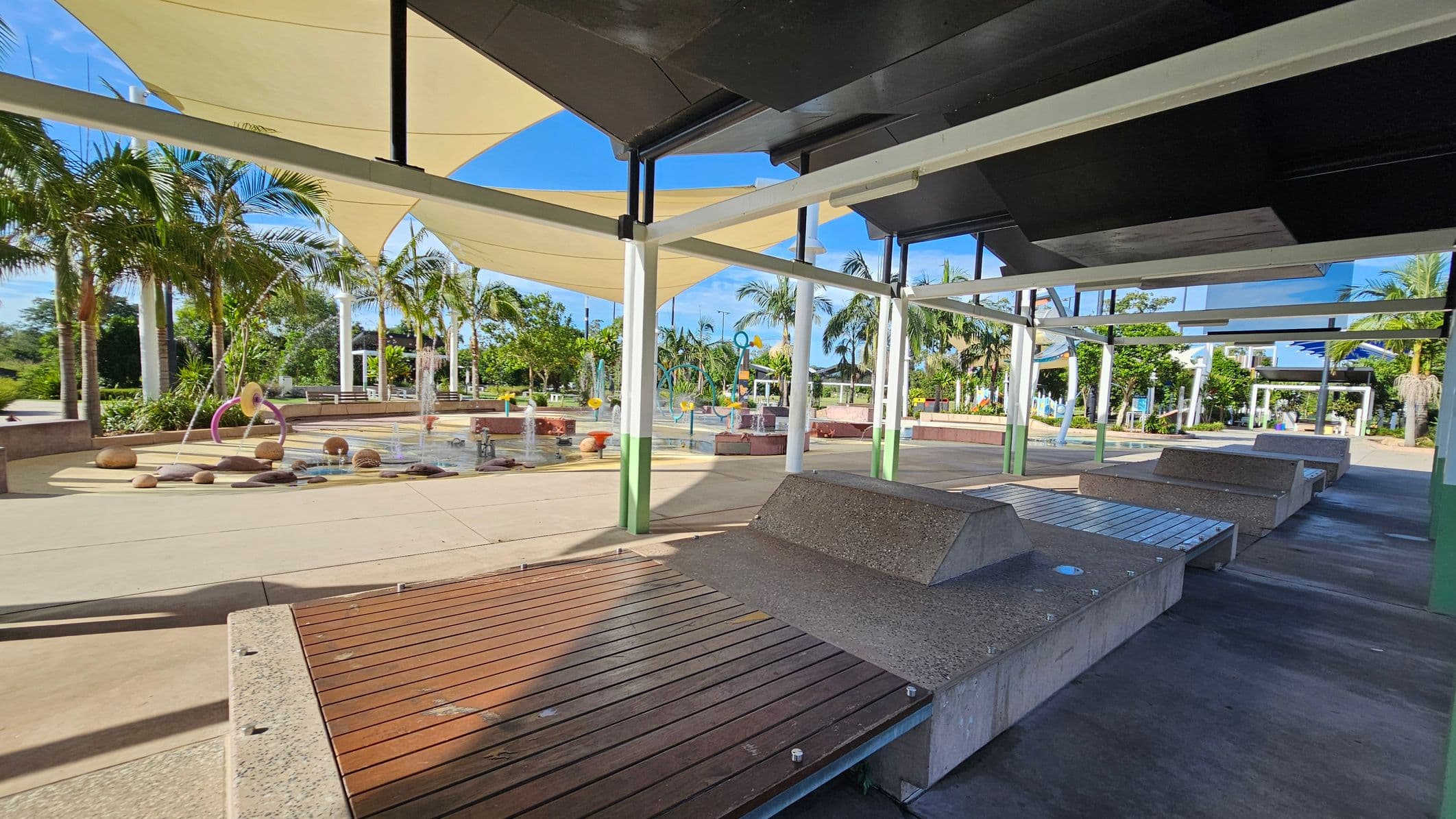 On the left, there are several palm trees and a water play area with fountains and colourful structures. The middle features a paved walkway with scattered benches and seating areas. On the right, there are shaded seating areas with wooden and concrete benches under a canopy. The background shows more greenery and additional park structures. The ceiling consists of large, overlapping fabric shades providing cover. The floor is a mix of concrete and paved surfaces.