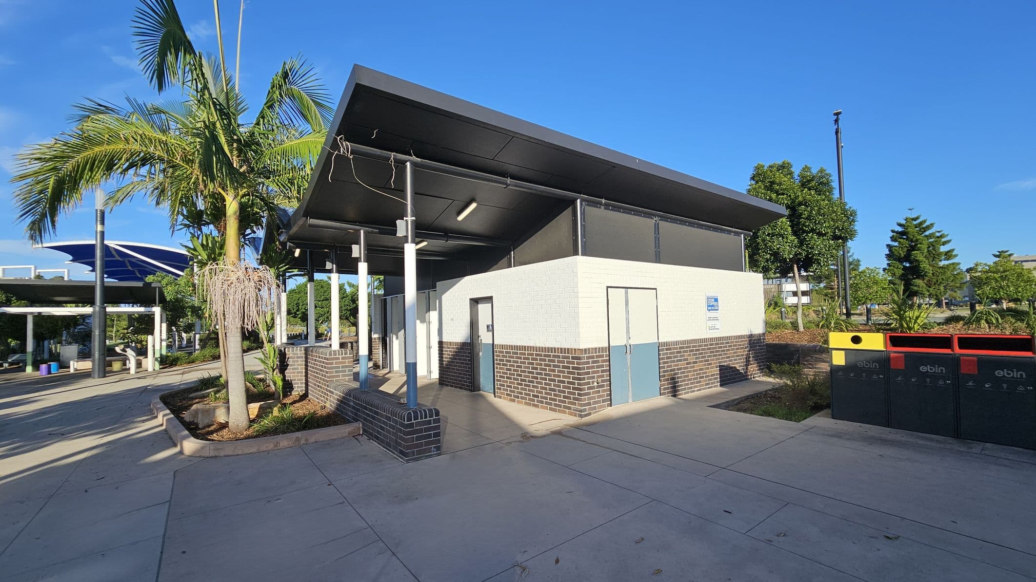 A public restroom building with a slanted roof is in the centre, featuring light-coloured walls and dark brick accents. On the left, a palm tree and a covered walkway with blue canopies are visible. On the right, there are three waste bins in yellow, red, and black. The ground is paved with light-coloured concrete, and there are trees and shrubs in the background under a clear blue sky.