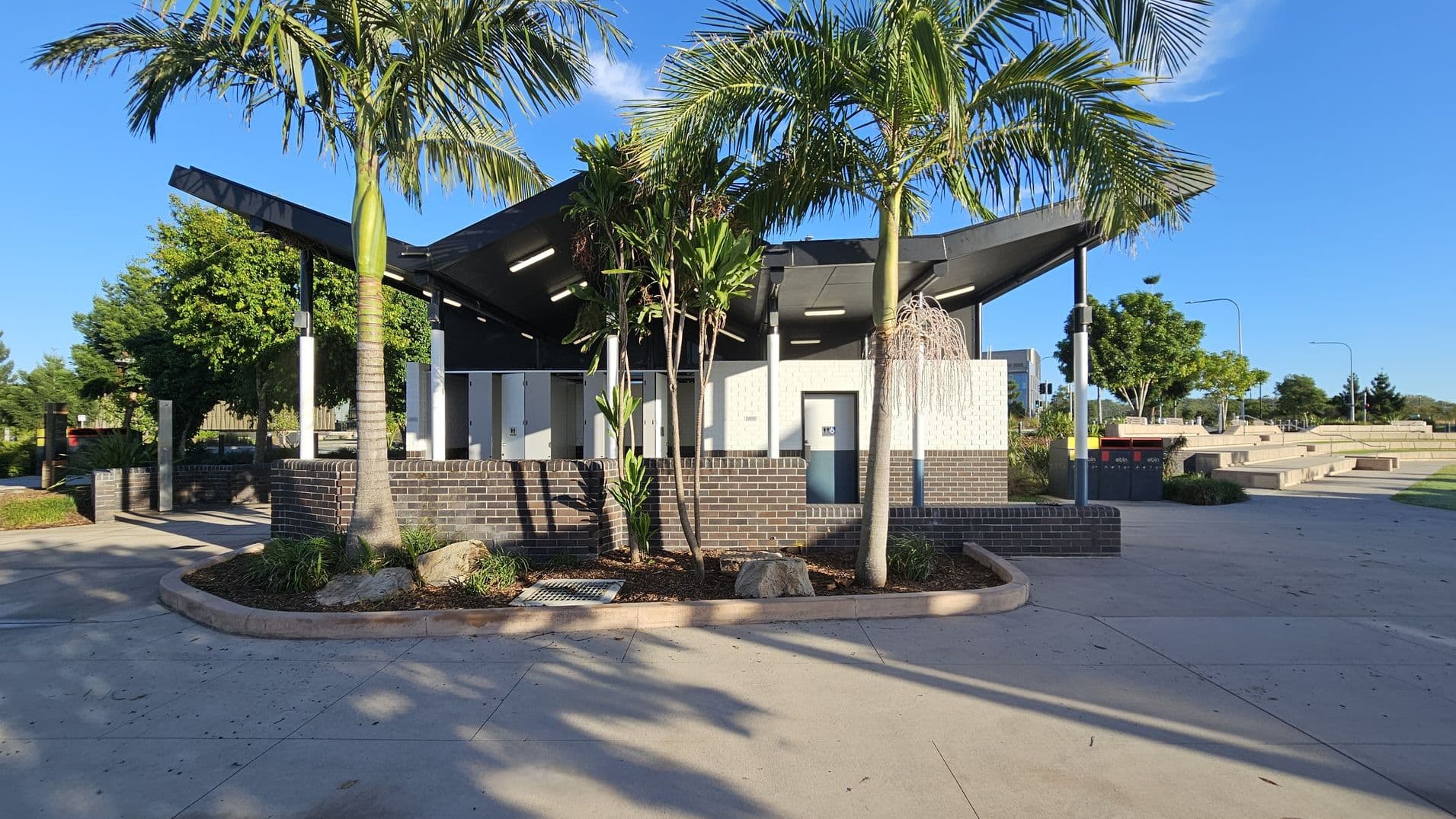 A public restroom facility with a modern design, featuring a black angular roof and white walls. In the foreground, there are several palm trees and small plants surrounded by a low brick wall. The ground is paved with light-coloured concrete. To the right, there are benches and a red bin. In the background, more trees and a clear blue sky are visible.