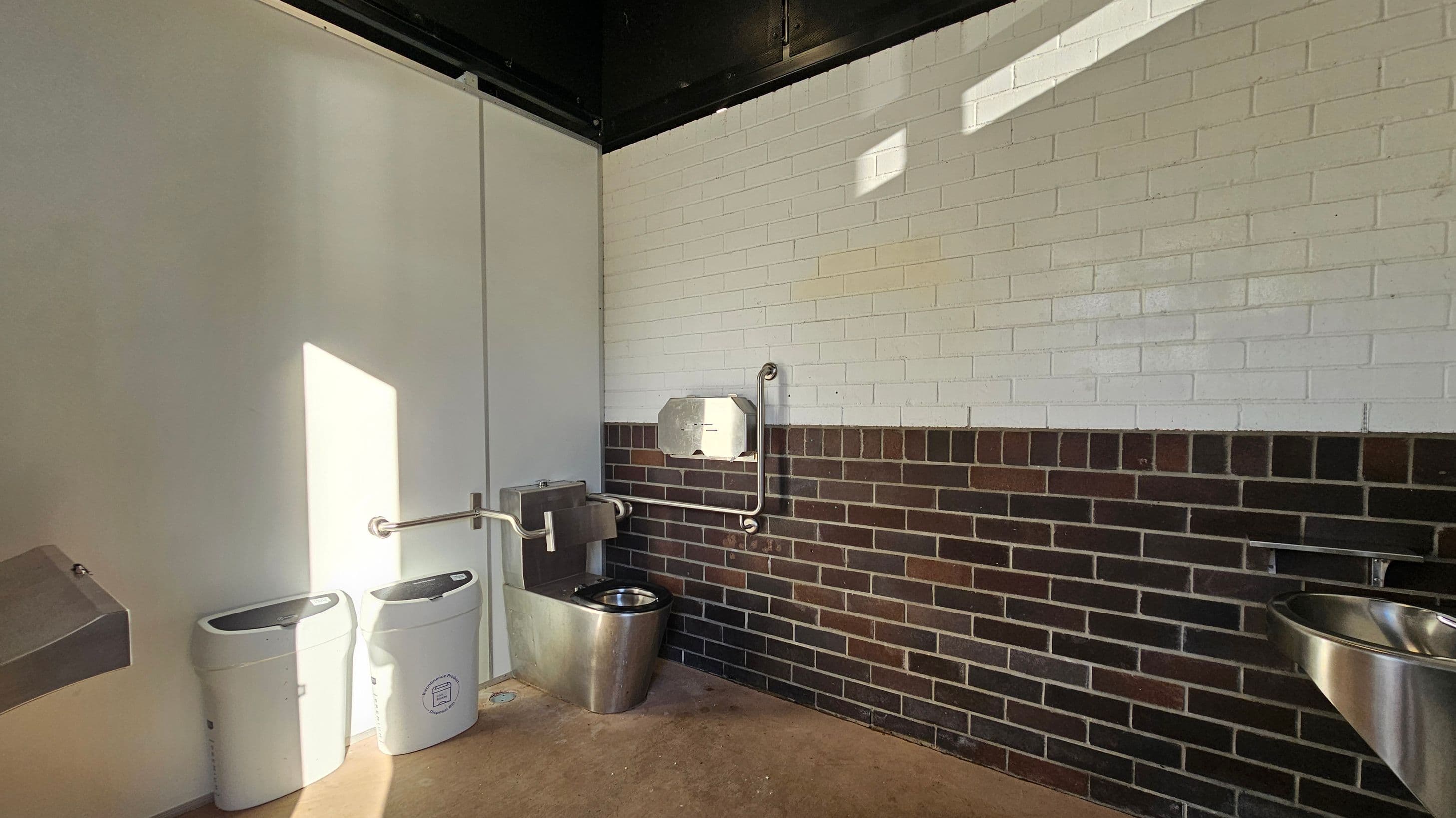 A public restroom with a stainless steel toilet and sink on the right. The walls are tiled with white bricks on the upper half and dark bricks on the lower half. On the left, there are two white bins beneath a metal hand dryer. The floor is concrete, and the ceiling is black. A metal handrail is mounted on the wall near the toilet.