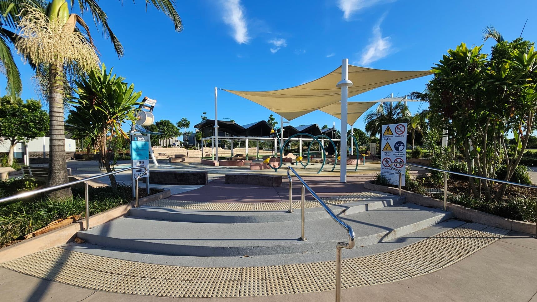 A playground area with a smooth, light-coloured concrete floor and a metal handrail leading up steps. In the centre, there are large shade sails supported by poles, covering play equipment. On the left, a palm tree and lush greenery are visible, along with a sign. On the right, more greenery and a sign with various warnings. In the background, there are additional trees and a building with a dark roof. The sky is clear and blue.