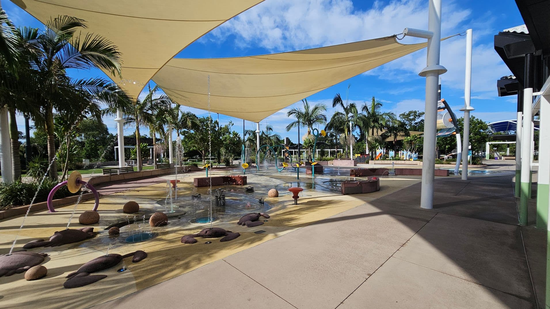 A water play area with various fountains and water features on a wet, textured surface. Overhead, large triangular shade sails provide cover. Palm trees and other greenery surround the area, with benches and pathways visible in the background. The ground is a mix of wet and dry concrete, and there are colourful play structures scattered throughout. White poles support the shade sails, and a building with a dark roof is partially visible on the right.
