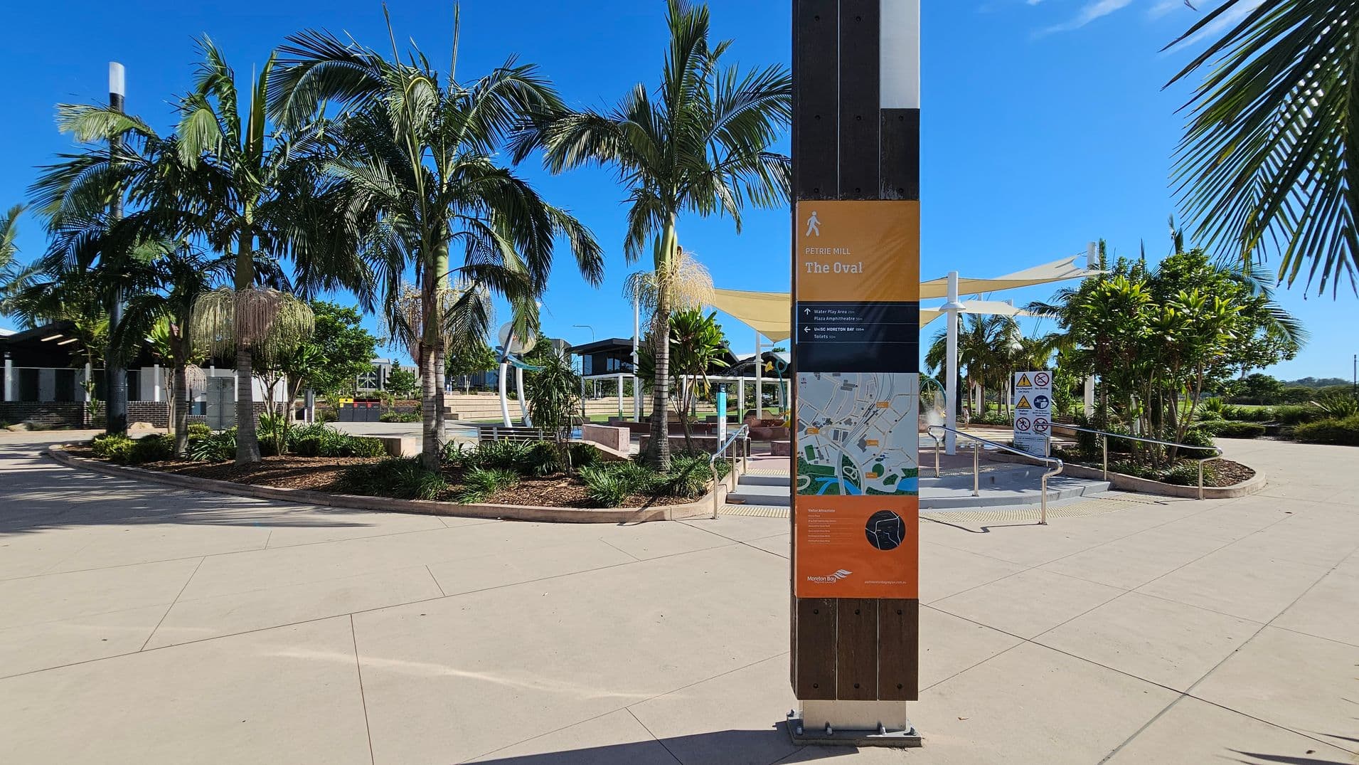 A public space with a paved walkway and a central signpost displaying a map and information about "The Oval." On the left, there are several tall palm trees and a building with a shaded area. In the middle, more palm trees and landscaped garden beds are visible. On the right, a path leads to a shaded area with white canopies and additional signage. The sky is clear and blue.