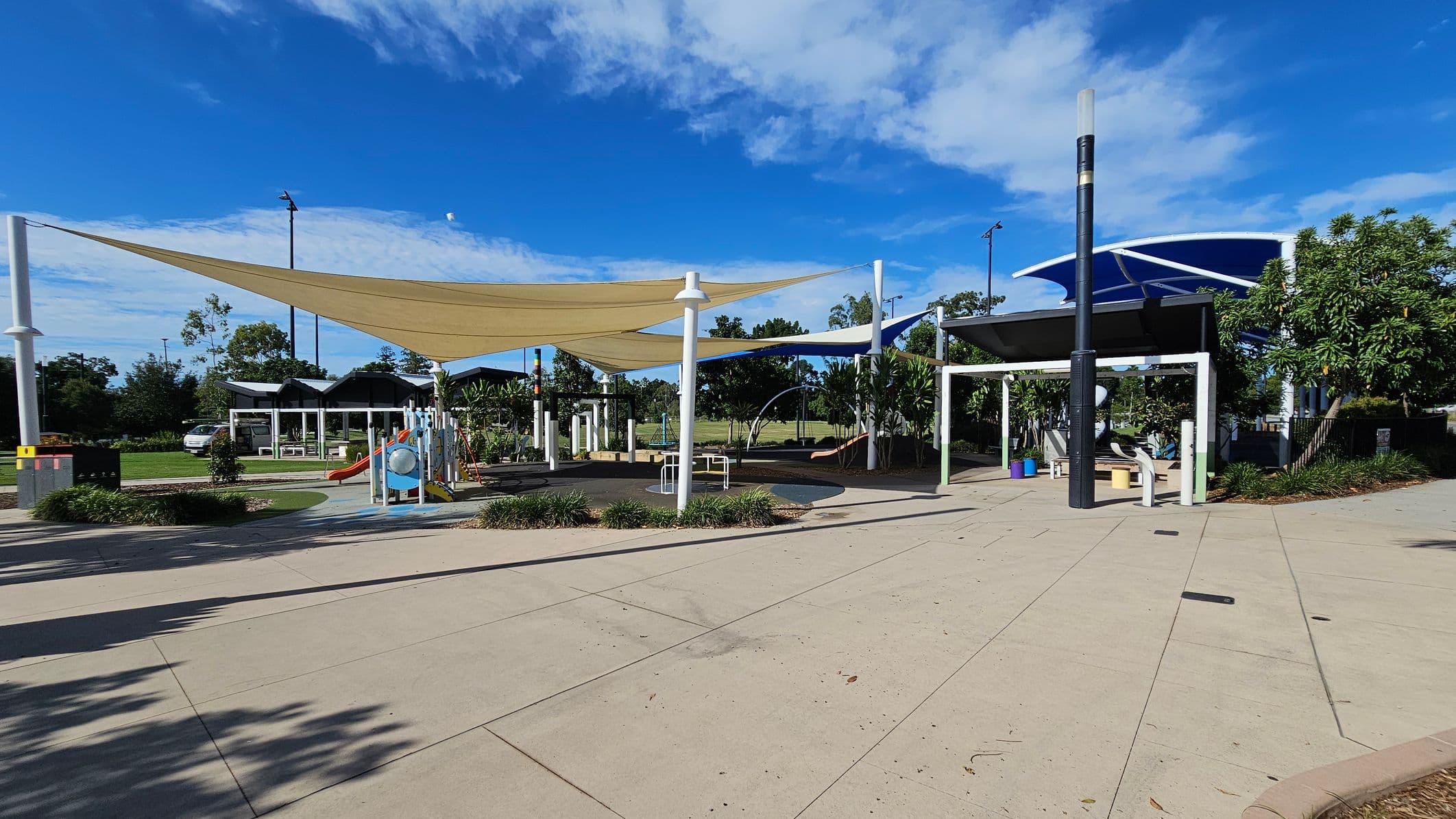 A playground with various equipment, including a slide and climbing structures, is covered by large shade sails. The ground is a mix of soft play surfaces and concrete paths. On the right, there is a sheltered seating area with a table and benches. Trees and shrubs are scattered throughout, providing greenery. In the background, a grassy field and more trees are visible. A car is parked on the left side near a small building or shelter.