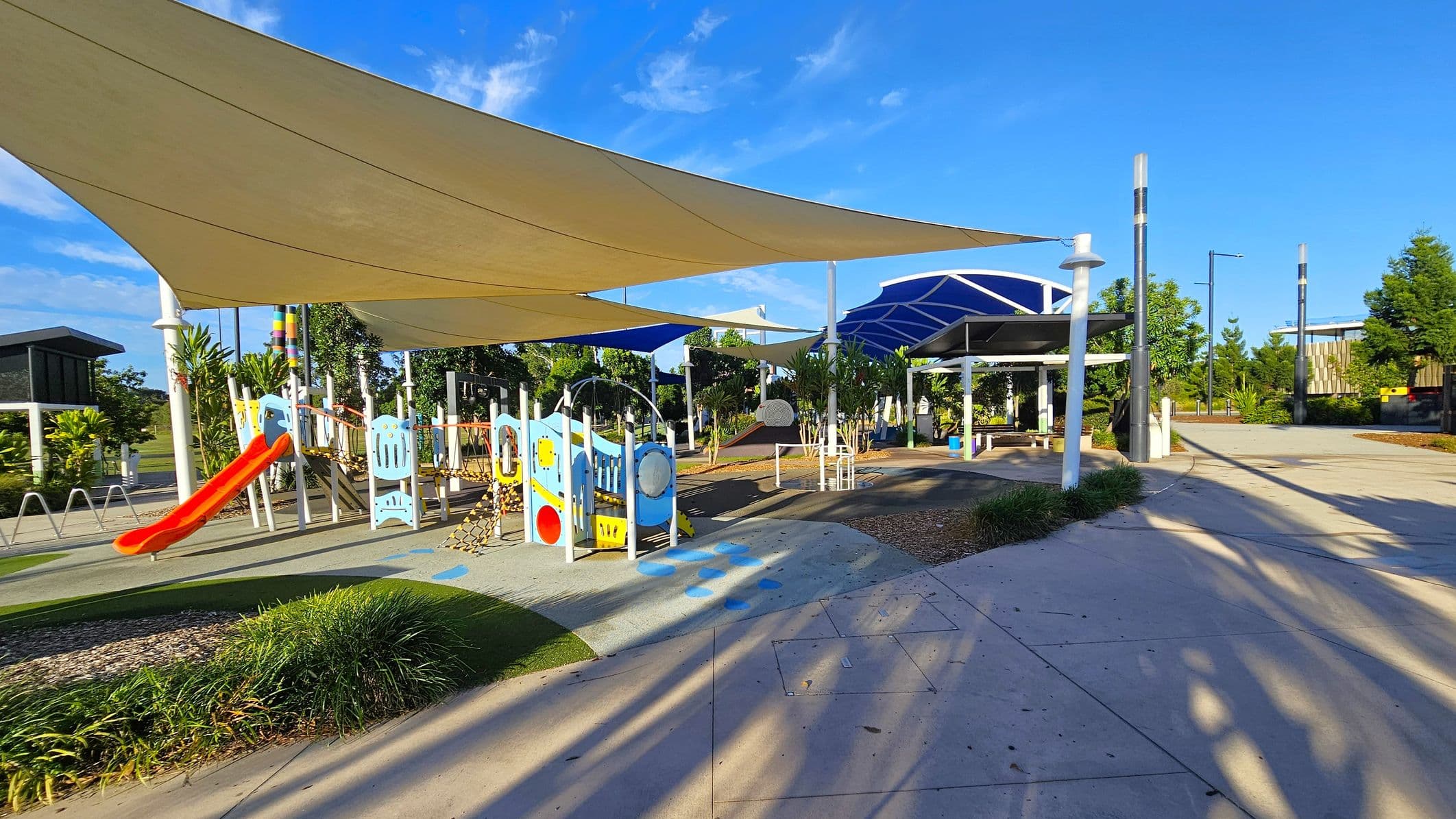 A playground with a red slide and colourful climbing structures on the left, shaded by large triangular sails. The ground is a mix of concrete and soft play surfaces. In the middle, there are small trees and plants. On the right, a covered seating area with benches and a pathway leading further into the park. The background features more trees and a building. Shadows from the sails and structures are cast on the ground.