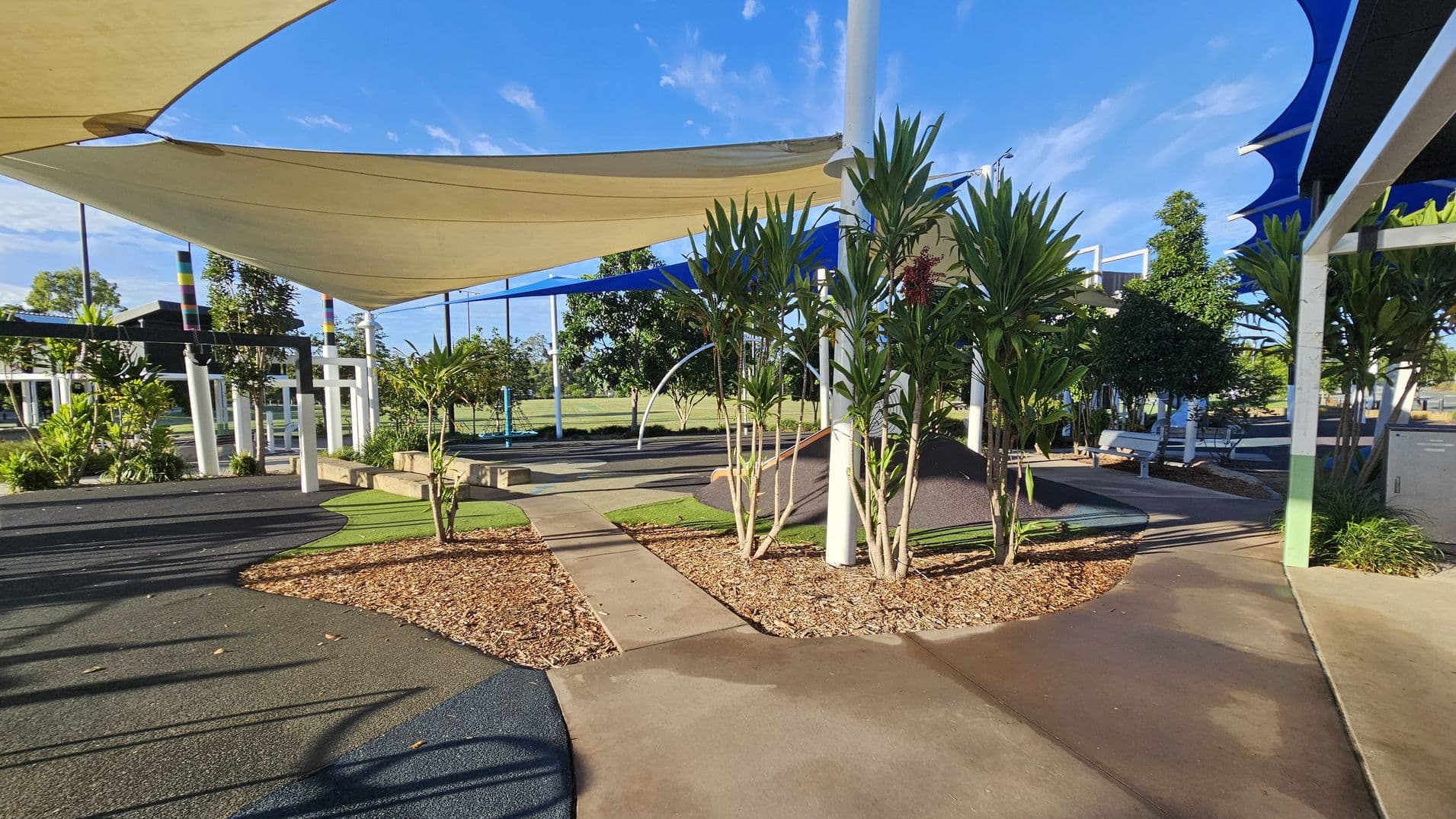 A playground with a mix of concrete and rubberised paths. In the centre, there are small trees and plants surrounded by mulch. Overhead, large triangular shade sails in beige and blue provide cover. To the left, colourful poles support the structure, and there are various play equipment pieces. In the background, more trees and a grassy field are visible. Benches are placed along the paths, and the area is well-maintained with a clean, modern design.