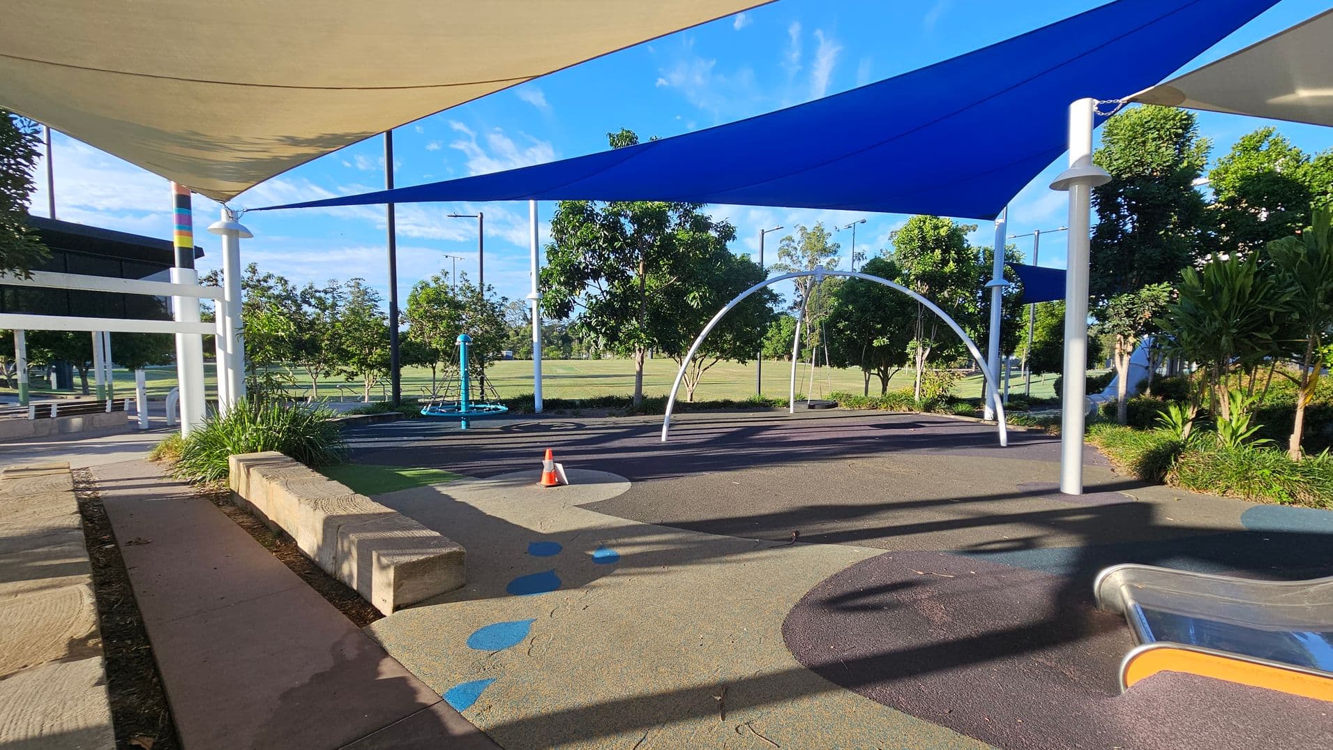 A playground area with colourful shade sails overhead, including blue and beige. On the left, there are concrete benches and a small orange traffic cone on the ground. In the centre, a metal arch structure and a blue climbing frame are visible. The ground is covered with a textured, multicoloured surface. Trees and shrubs are scattered throughout the background, with a grassy field beyond.