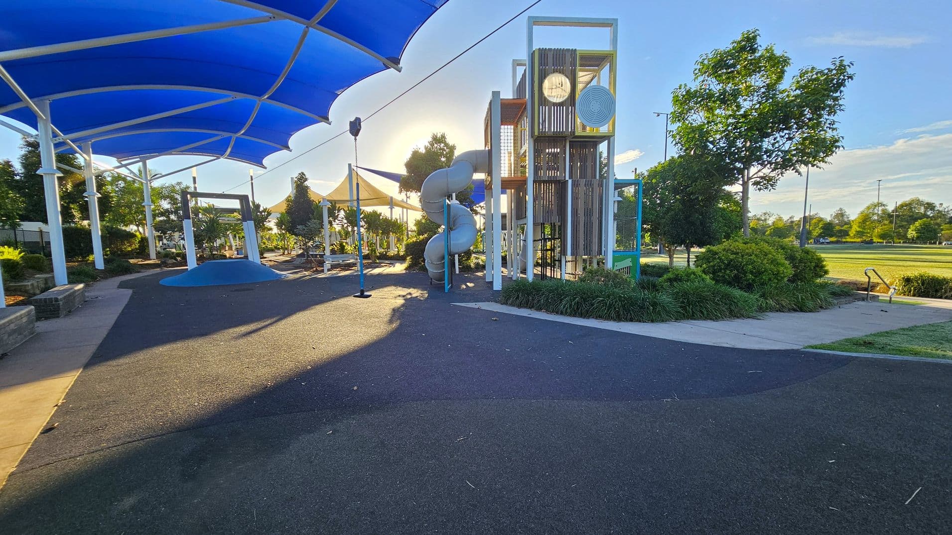 A playground with a large blue shade structure on the left, covering a play area with a small mound and a swing. In the centre, there's a tall play structure with a spiral slide. The ground is covered with a dark rubber surface. On the right, there are trees and bushes along a concrete path leading to an open grassy field. The sun is low, casting long shadows across the playground.