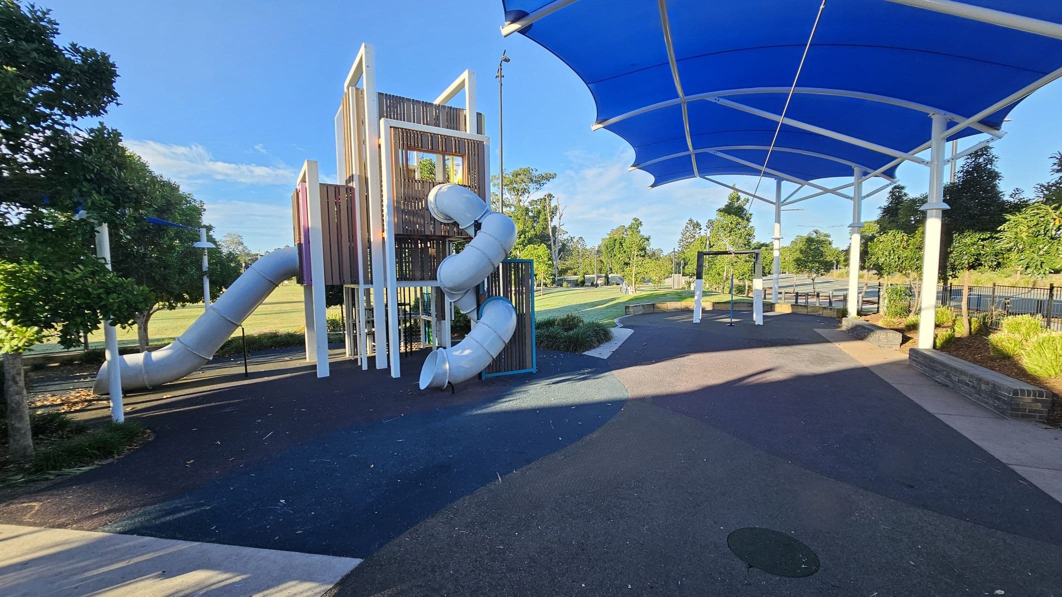 A playground with a large, multi-level climbing structure on the left, featuring a twisting tube slide. The ground is covered with a dark, rubberised surface. On the right, there is a blue shade canopy supported by white poles, providing cover over part of the play area. In the background, there are trees and a grassy field. A path runs alongside the playground, bordered by low brick walls and landscaped greenery.