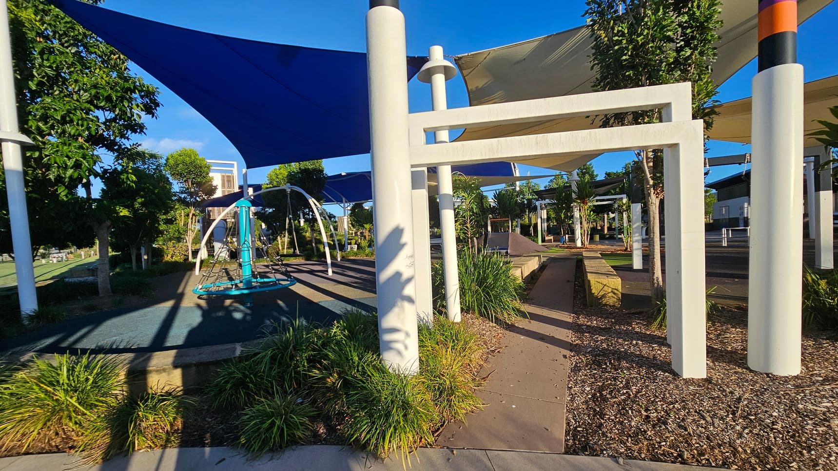 A playground with large white poles supporting blue and beige shade sails. On the left, there's a circular swing structure with a blue base. The ground is covered with a mix of concrete, mulch, and grass. Trees and shrubs are scattered throughout, providing greenery. In the background, there are more playground structures and a building partially visible. Shadows from the structures and trees are cast on the ground.