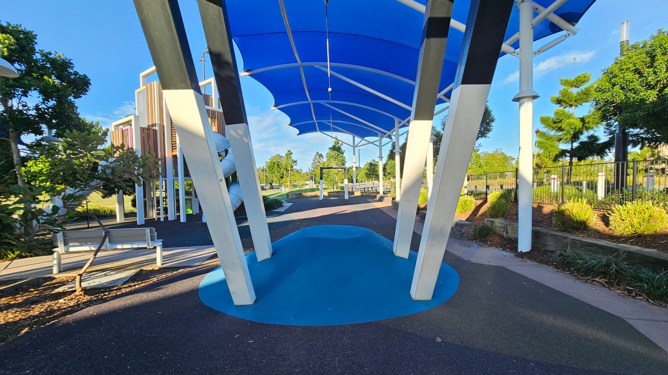 A playground with a blue canopy overhead supported by white beams. In the foreground, there's a blue mound on the ground. To the left, a white bench is near some trees and a play structure with slides. On the right, a fence runs along the edge with more trees and shrubs. The ground is a mix of black and blue surfaces.