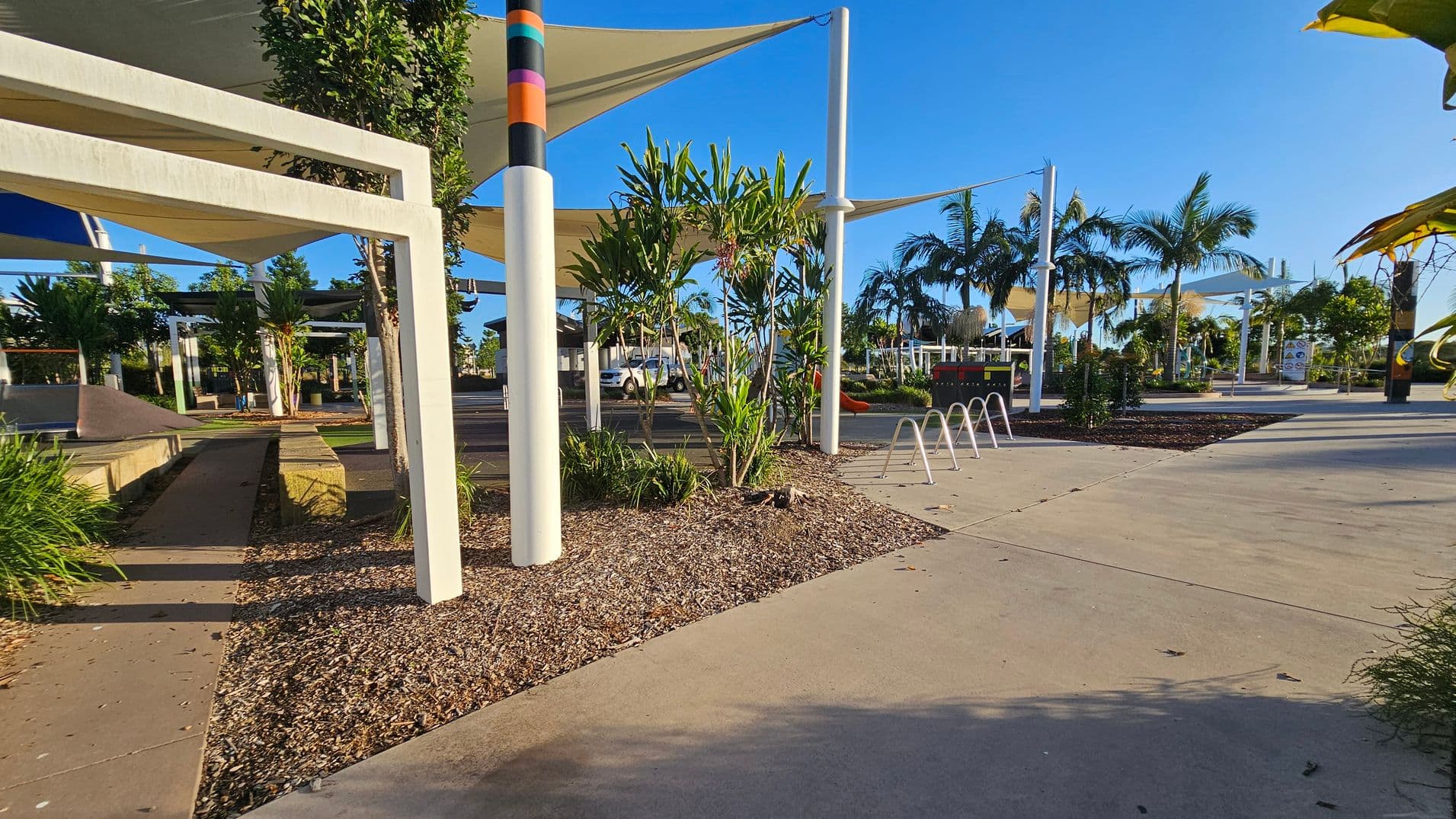 A public park area with a concrete path and several white metal structures providing shade. On the left, there are raised garden beds with green plants and a small ramp. In the centre, a colourful pole and a bike rack are visible. The right side features more plants and palm trees, with a playground in the distance. The ground is a mix of concrete and mulch, and the area is well-lit with clear skies.