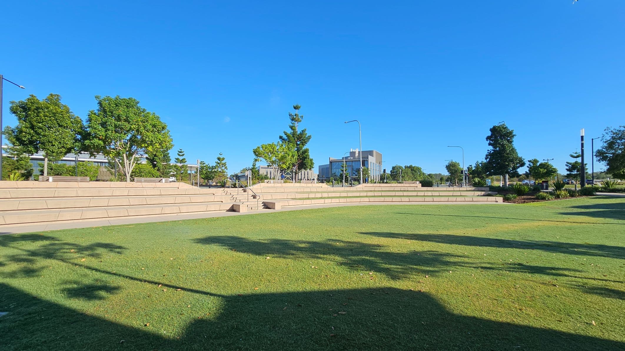 An open grassy area with tiered stone seating in the foreground. In the background, there are several trees and a modern building with large windows. The area is well-lit with street lamps, and there are pathways and benches visible. The sky is clear and blue.