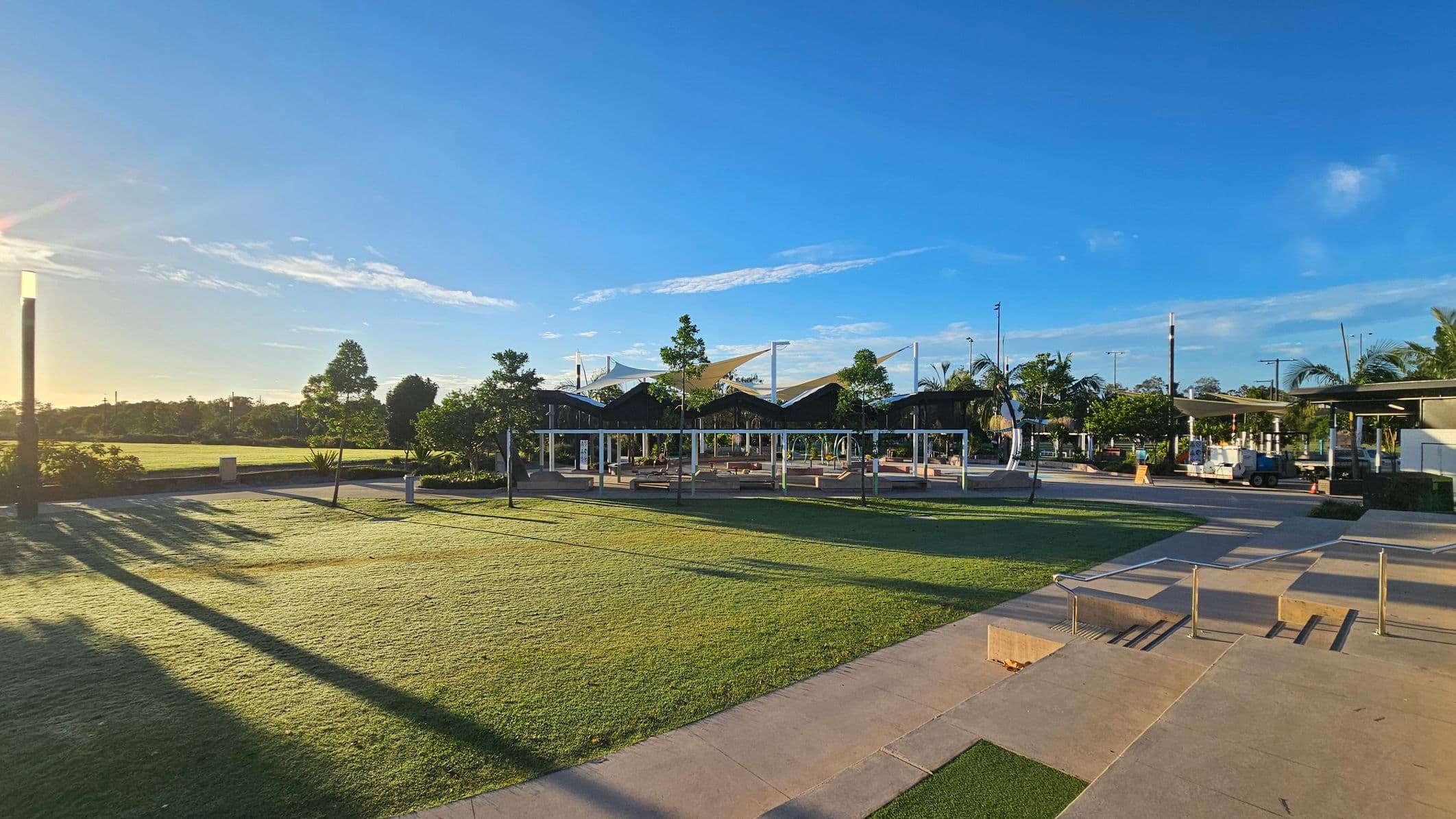 A park with a grassy area in the foreground, casting long shadows from the trees and structures. In the middle, there are several modern, open-air shelters with angular roofs and white support beams. Trees and shrubs are scattered throughout the area. On the right, a paved walkway with steps and metal railings leads towards the shelters. The background features more trees and a clear blue sky.
