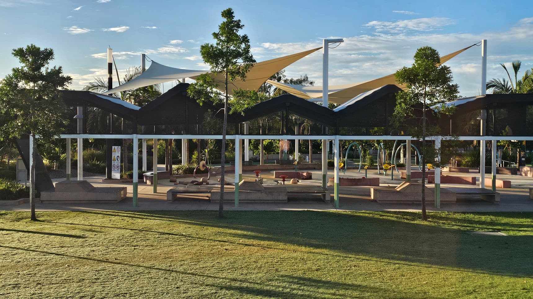 A park area with a grassy foreground and a playground in the background. The playground features various play equipment, including swings and climbing structures. There are several trees scattered throughout the area, providing shade. Overhead, there are large, triangular shade sails supported by poles. The ground is a mix of grass and paved surfaces, and there are benches and seating areas under the shade structures.