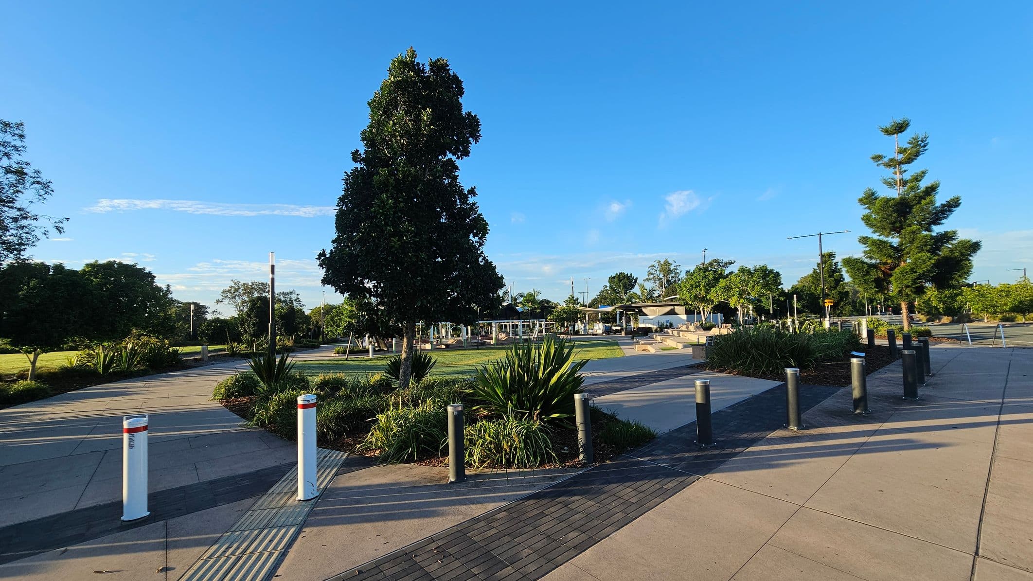 A park scene with a paved walkway in the foreground, bordered by white and black bollards. The path is lined with green shrubs and trees. In the centre, a large tree stands prominently, surrounded by grass. To the right, more trees and a structure with a roof are visible. The area is well-maintained, with clear paths and landscaped greenery. The sky is clear and blue.