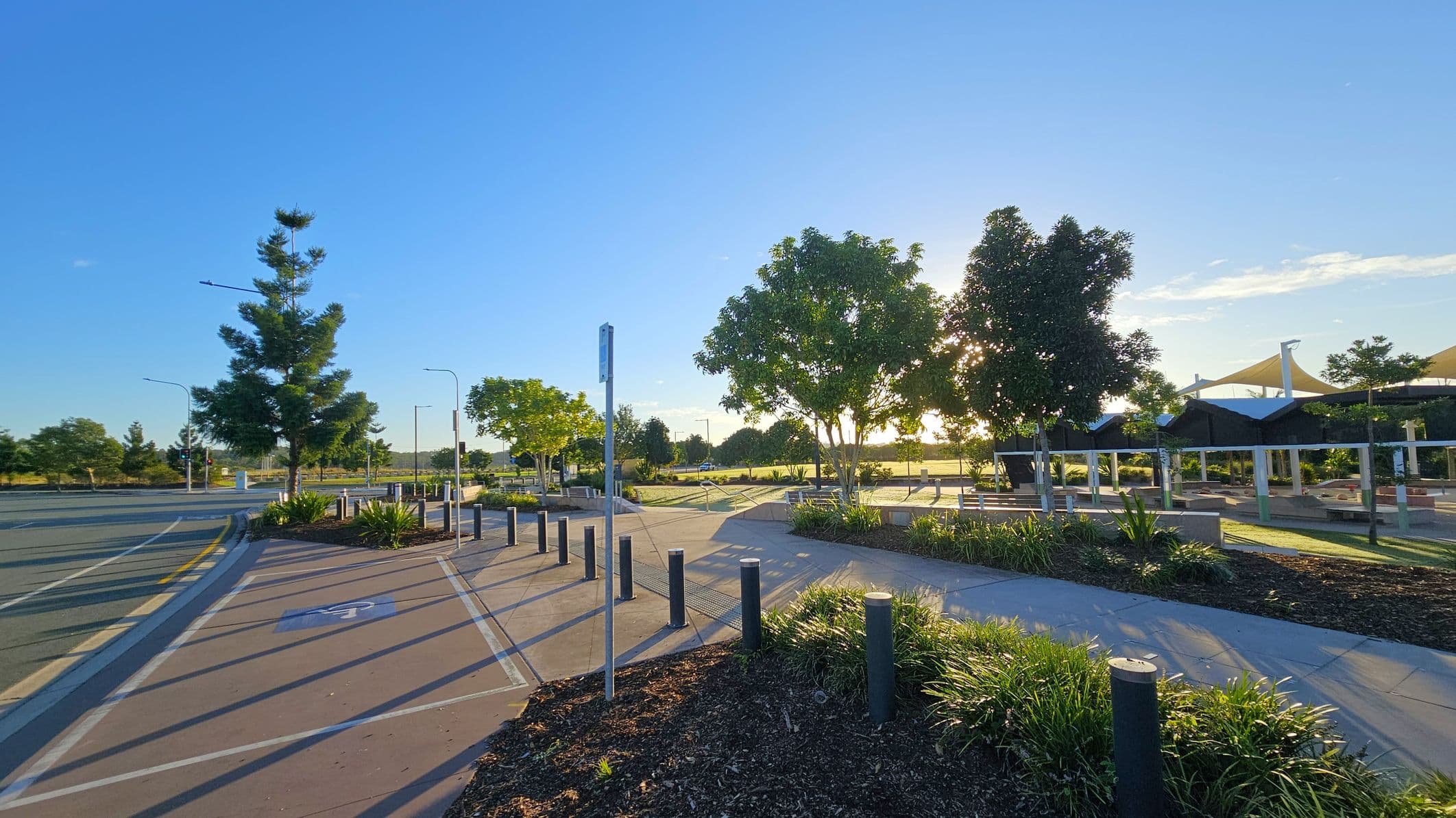 A paved pathway with a bicycle lane runs through the foreground, bordered by low black bollards and green shrubs. On the left, a tall tree stands near the road, which curves gently. In the centre, several trees provide shade, with sunlight filtering through. To the right, there are covered seating areas with triangular shade structures. The background features more trees and open grassy areas, with a clear blue sky above.