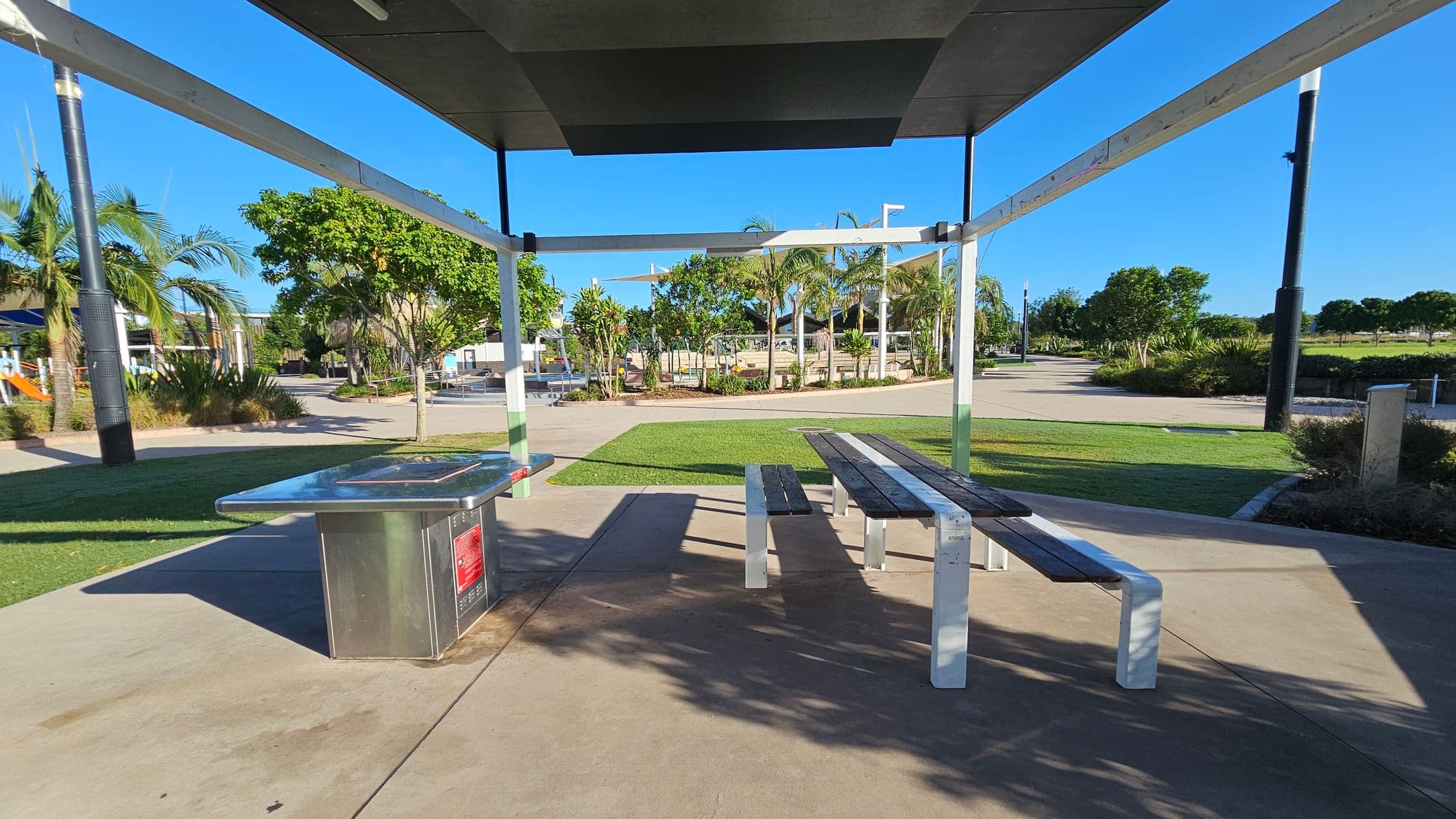 A park setting with a metal barbecue grill on the left and a wooden picnic table with benches on the right. The area is covered by a modern, angular shelter. In the background, there are palm trees and other greenery, with a paved walkway winding through the park. The ground is a mix of concrete and grass, and there are tall black poles scattered around the area.