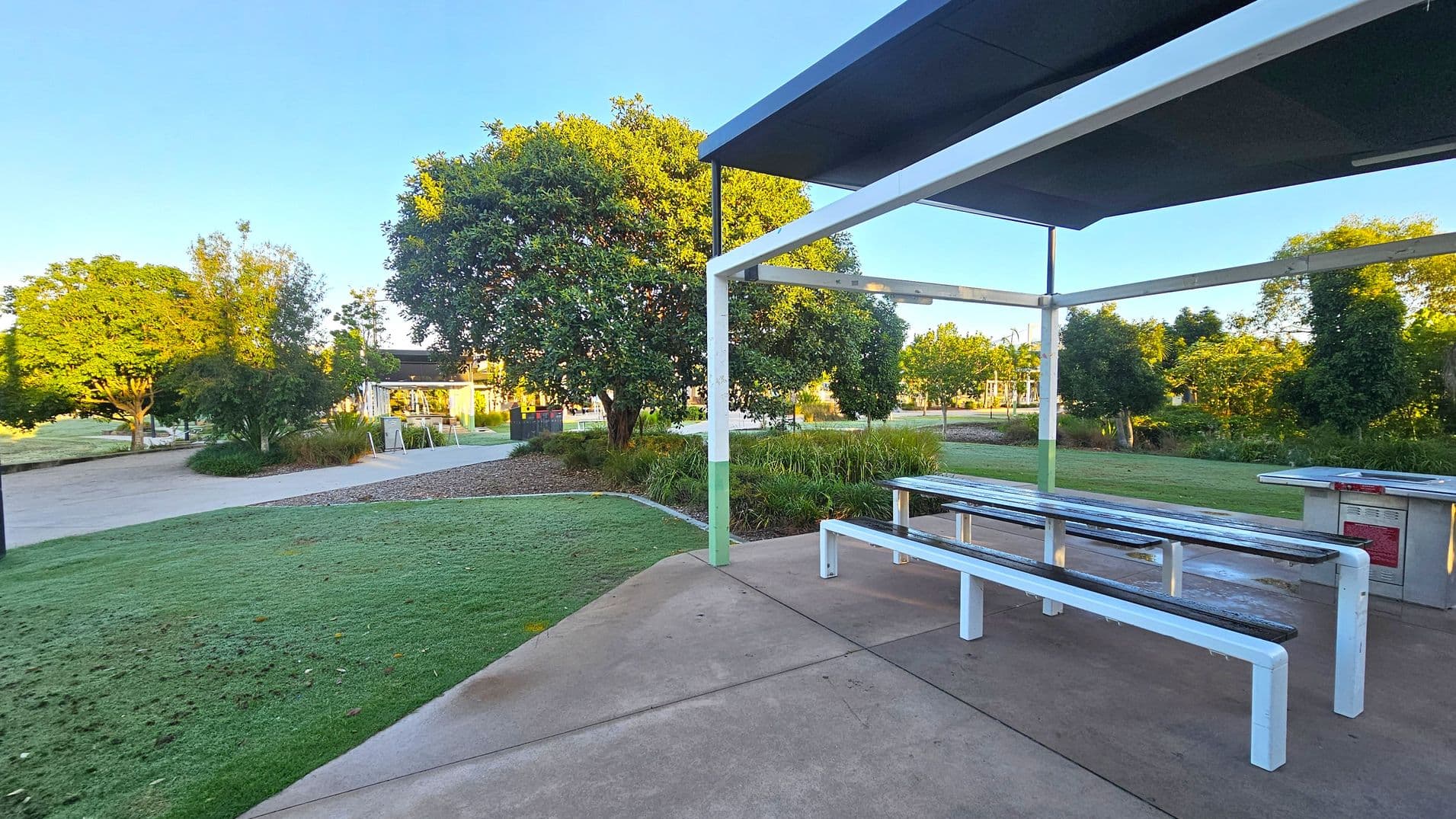 A park setting with a covered picnic area on the right, featuring a metal table and benches. The ground is paved with concrete. In the background, there are several trees and a grassy area. A pathway curves through the park, leading to a structure with a roof and open sides. The sky is clear, and the sunlight casts shadows on the ground.