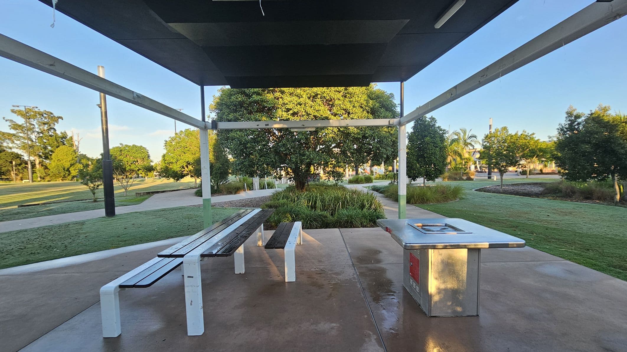 A park setting with a covered picnic area. In the foreground, there is a metal barbecue grill on the right and a picnic table with benches on the left. The ground is paved with concrete. In the background, there are well-maintained grassy areas and a variety of trees, including a large tree directly behind the picnic area. The structure above provides shade, supported by metal beams. Paths wind through the park, and there are more trees and shrubs in the distance.