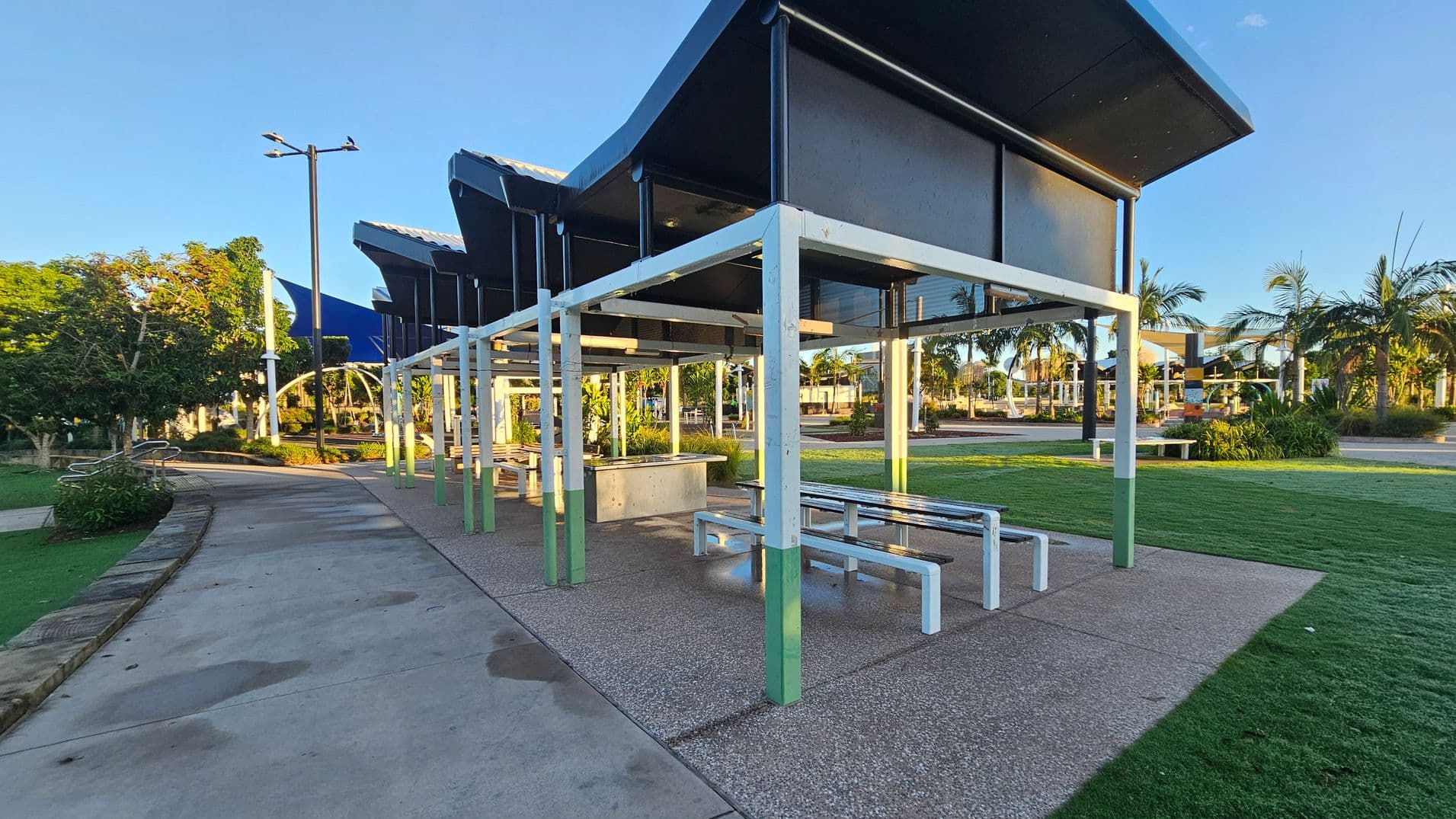 A park setting with a covered seating area in the foreground, featuring white metal frames and benches. The ground is a mix of concrete and grass. To the left, a paved pathway curves through the park, lined with trees and a lamp post. In the background, more trees and a structure with a blue roof are visible, along with additional seating areas and palm trees. The sky is clear and blue.