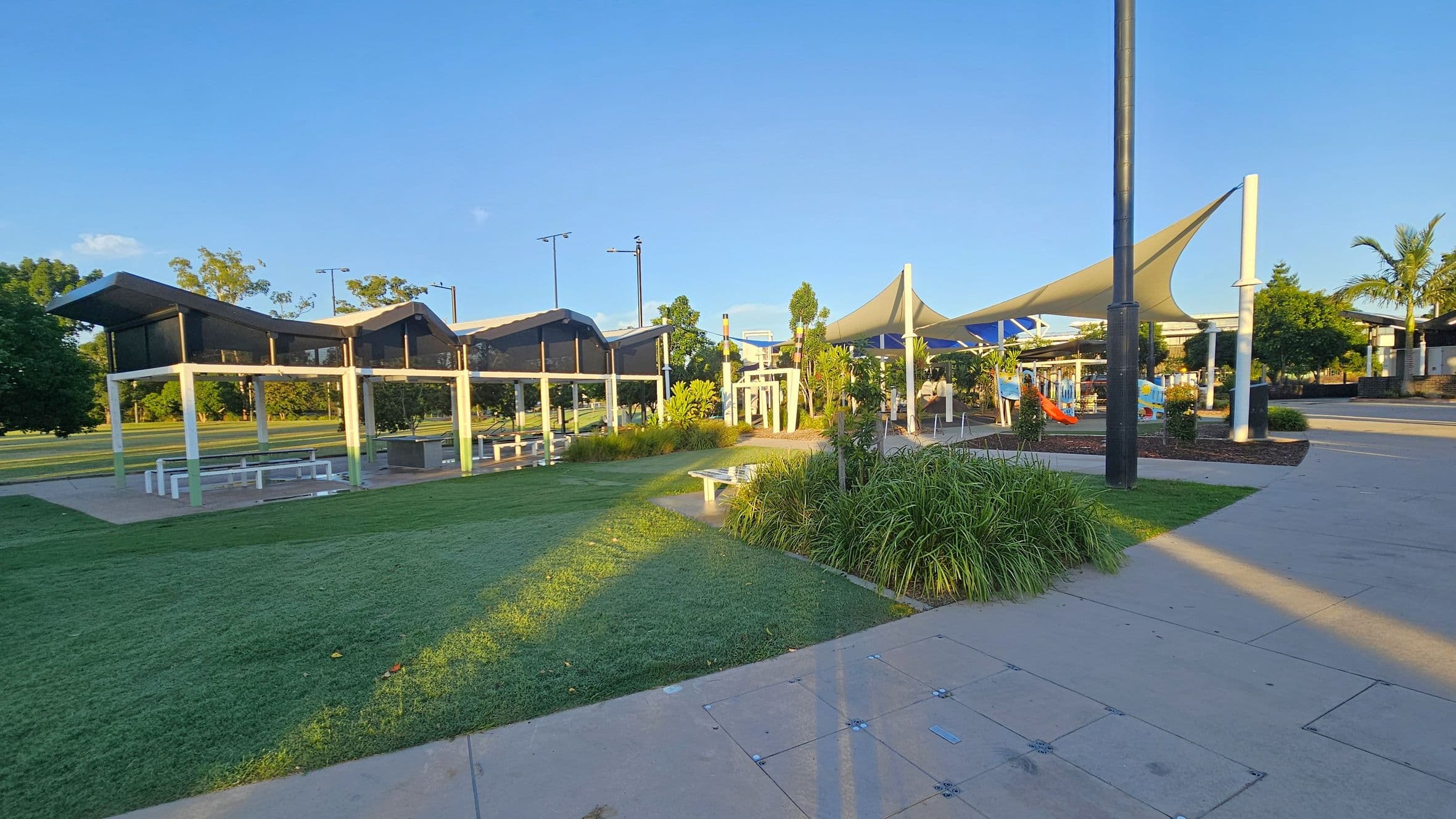 A park scene with a modern shelter on the left featuring a slanted roof and white pillars, covering picnic tables. In the centre, there are green lawns and landscaped plants. On the right, a playground with colourful slides is shaded by large triangular fabric canopies. A wide concrete path runs through the area, and a tall black pole stands near the playground. Trees and more greenery are visible in the background.
