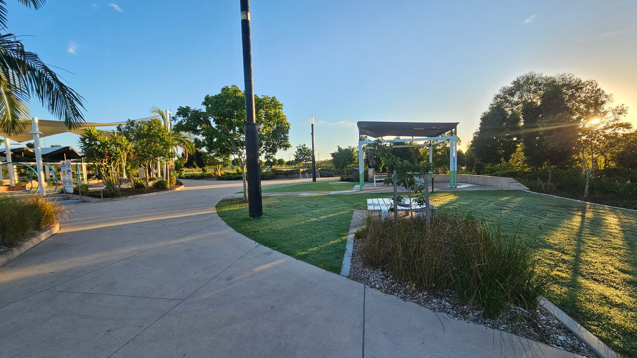 A park with a paved walkway curving through the scene. On the left, there are palm trees and a shaded area with white structures. In the centre, a grassy area with a small tree and a white bench. On the right, more trees with the sun setting behind them, casting long shadows. The foreground has a mix of grass and concrete, while the background features more greenery and a clear sky.