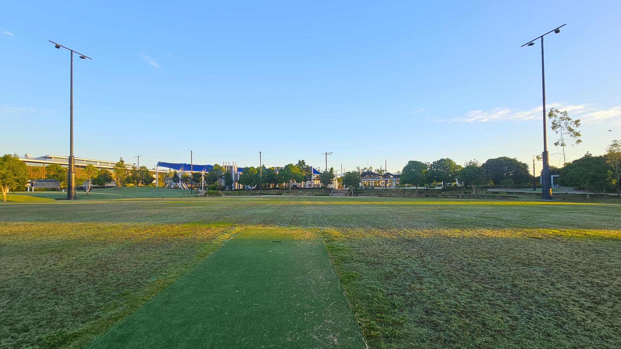 A grassy field with a green cricket pitch in the centre. Tall floodlights are positioned on both the left and right sides. In the background, there are trees and a few buildings with blue roofs. The sky is clear, and the area is well-lit by natural light.