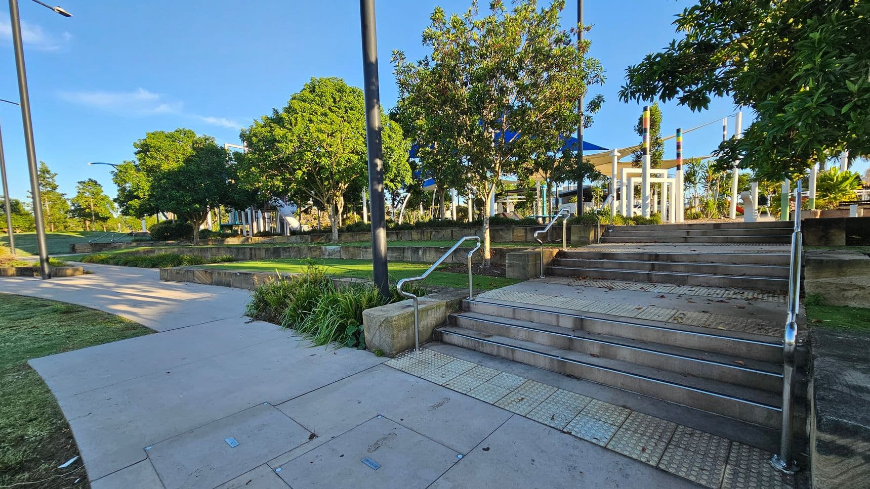 A park scene with a paved pathway on the left leading through a grassy area. On the right, a set of steps with metal railings ascends to a higher level. Trees with dense green foliage are scattered throughout, providing shade. In the background, there are white structures and a blue canopy, possibly part of a playground or seating area. The area is well-maintained, with neatly trimmed grass and landscaped plants.
