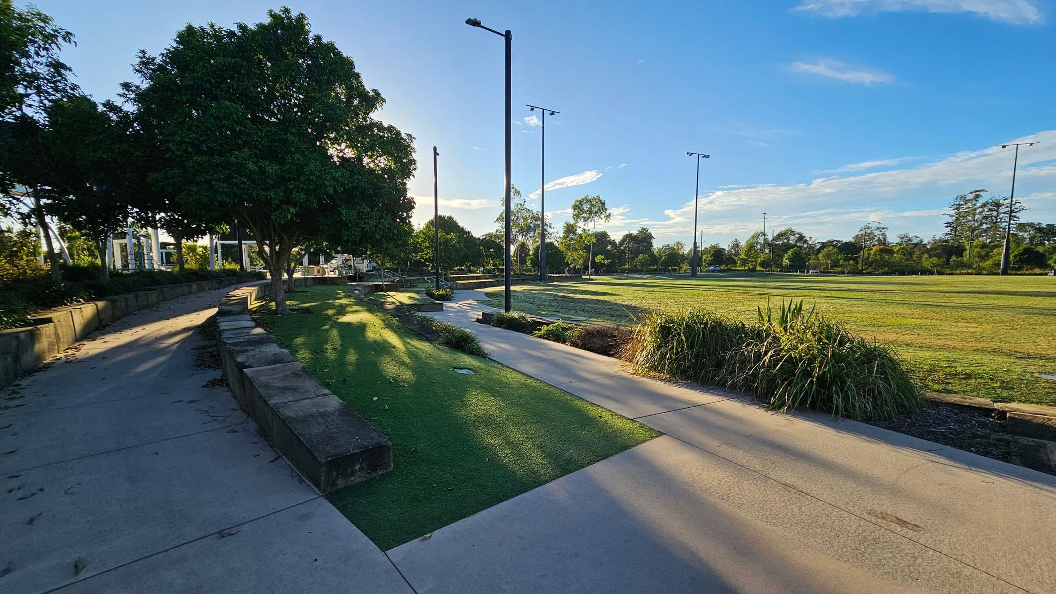 A park scene with a concrete path curving to the left, bordered by a low stone wall. On the left, there are large trees providing shade. To the right, an open grassy field is visible, with scattered tall light poles. Bushes and small plants line the path. The sky is clear with a few clouds, and the sunlight casts long shadows on the ground.