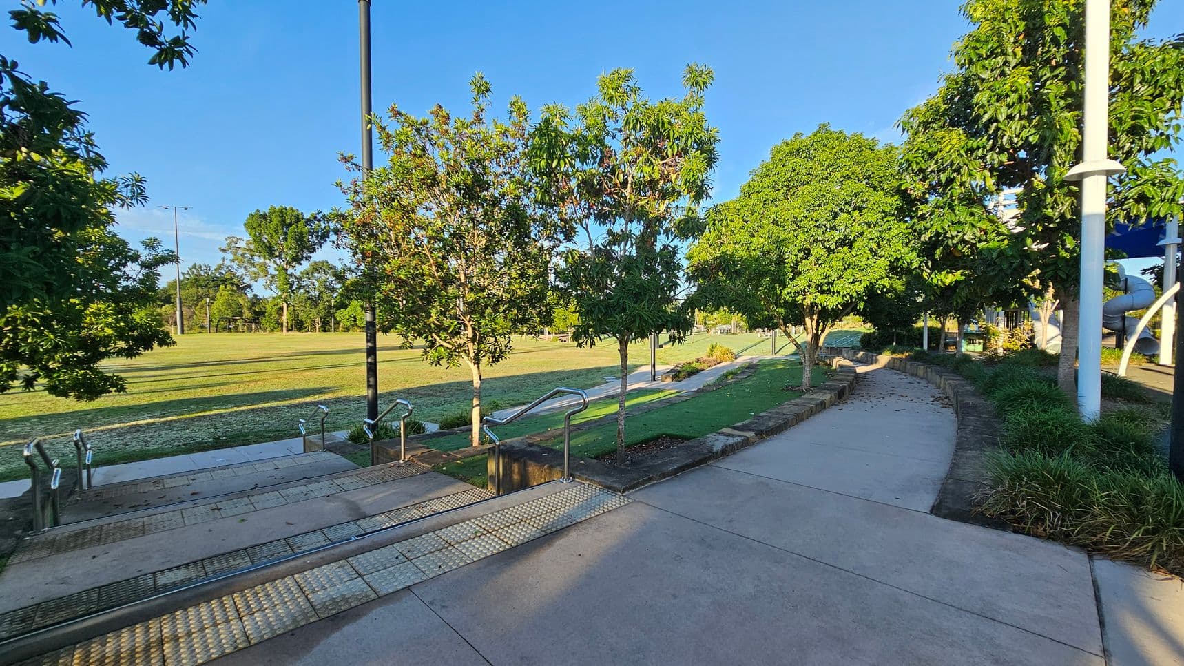 Concrete pathways and steps with metal railings in the foreground, leading down to a grassy area. Trees line both sides of the path, providing shade. On the right, a curved path is bordered by low stone walls and bushes. In the background, a large open field is visible, with more trees and a tall light pole. A playground structure with a slide is partially visible on the far right. The area is well-maintained and bathed in sunlight.
