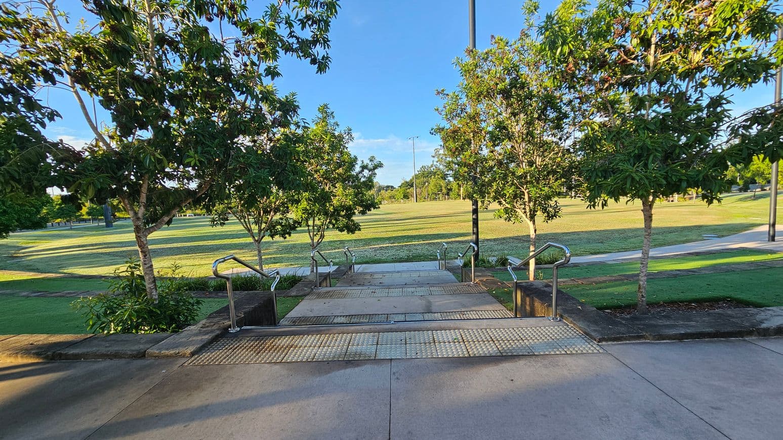 Concrete steps with metal railings lead down to a grassy field. Trees with green foliage line both sides of the steps, casting shadows on the ground. The field in the background is expansive, with a few scattered trees and a pathway running through it. The sky is clear and blue.