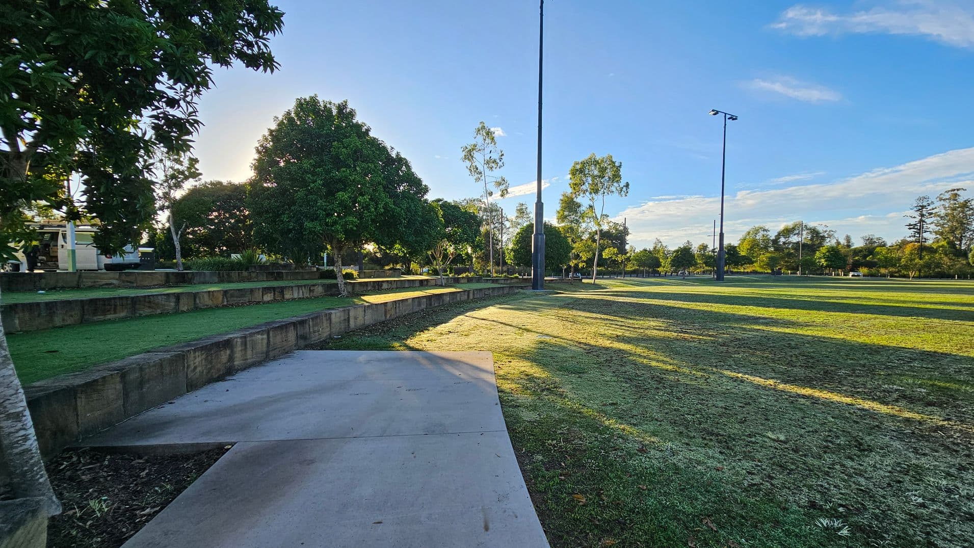 A park scene with a concrete path in the foreground leading to a grassy area. On the left, there are tiered stone seating areas and several large trees providing shade. In the middle, a tall light pole stands prominently. The background features more trees and open grassy fields, with long shadows cast by the setting sun. The sky is clear with a few clouds.