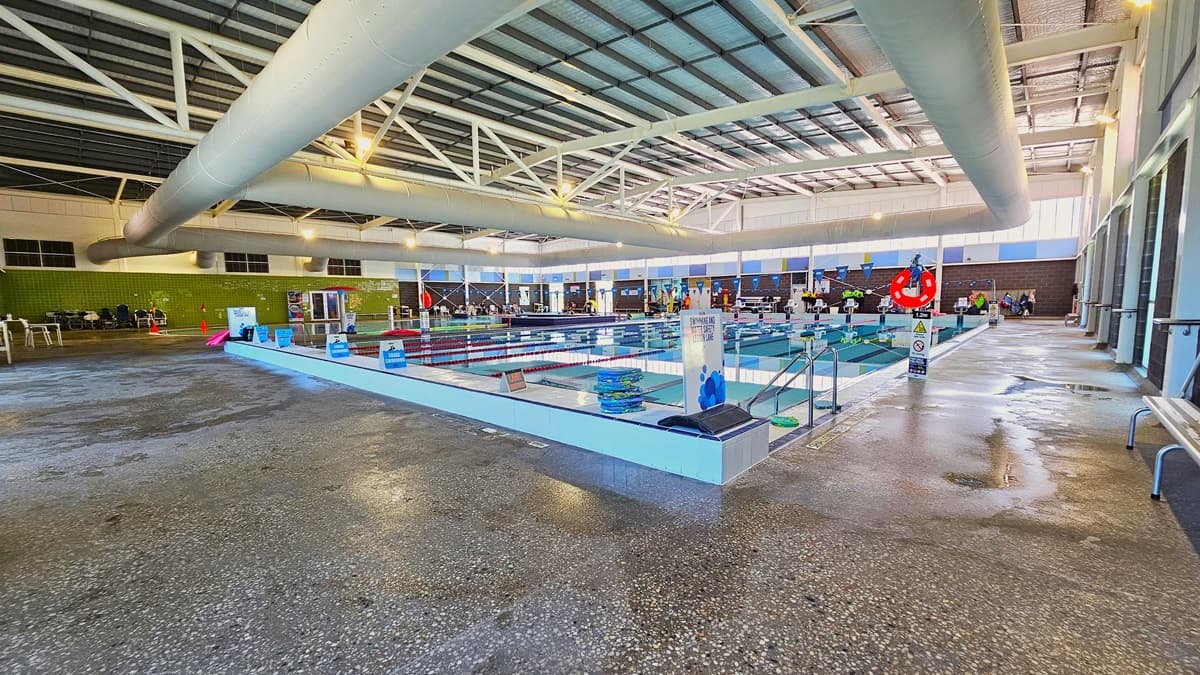 The 8-lane, 25m swimming pool has handrails in the corner, with swim accessories like red noodle hoses and floaters on the pool deck. On the right, there is bench seating, and on the left, there are aquatic wheelchairs aligned against the green tiled wall. The floor is non-slip concrete, and safety reminders, including blue triangle banners, are displayed. The high ceiling features insulation foams, large air-conditioning vents, metal beams, and bright lights.