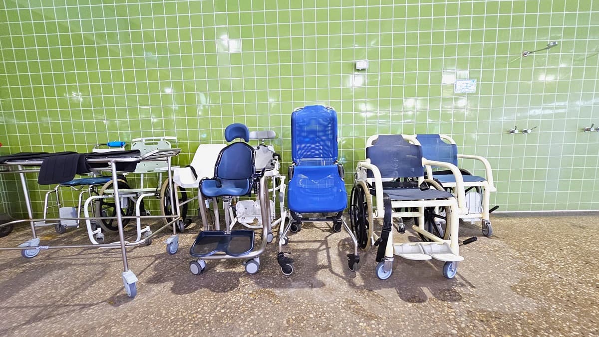 Aquatic wheelchairs and a cart with towels are positioned against the green-tiled wall, adjacent to the showers. The floor is made of non-slip concrete.