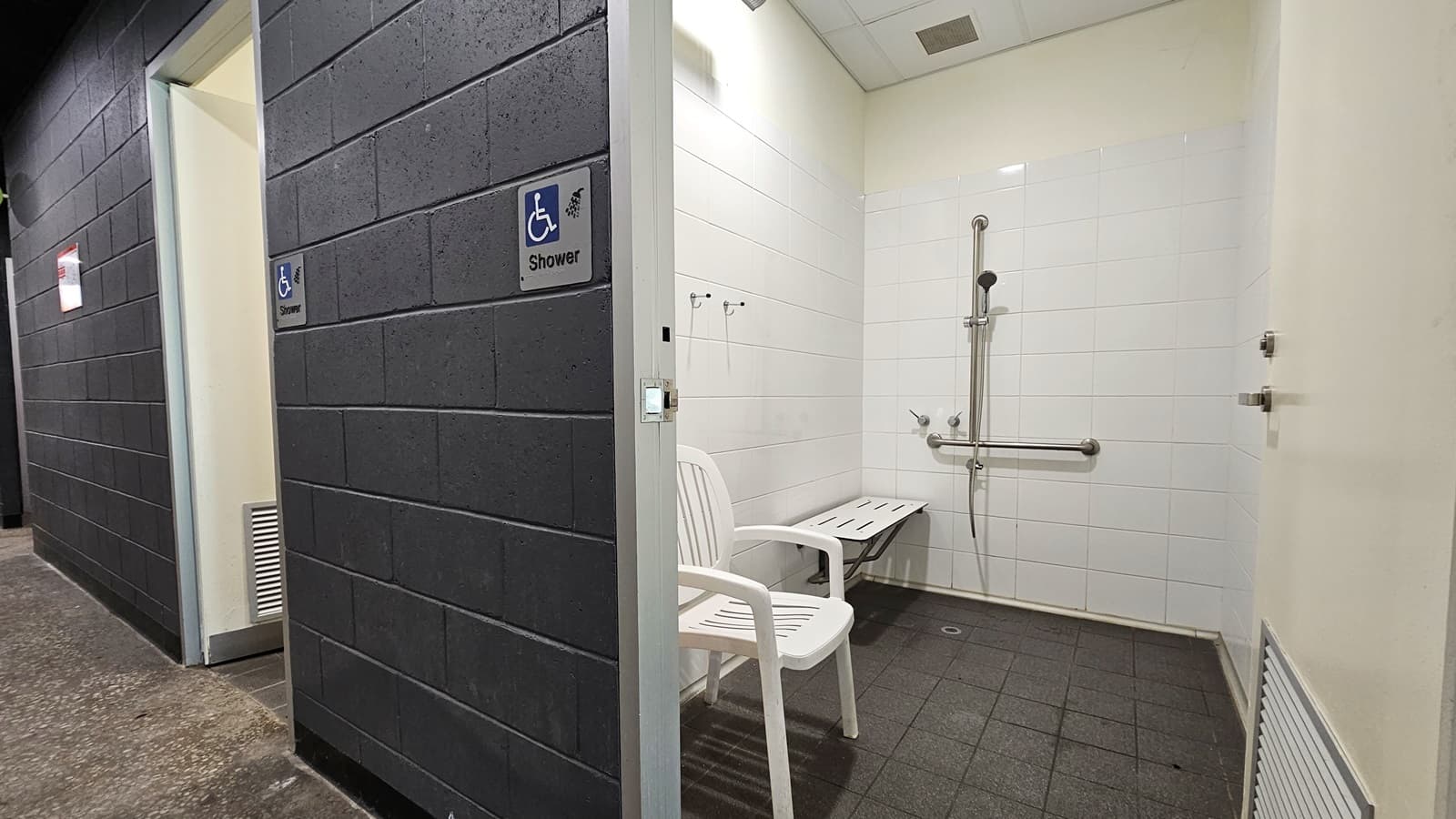 The shower facilities feature a sign indicating "Accessible Shower" on the dark grey brick wall. Inside, there is a shower with handrails, accompanied by a foldable bench and a white plastic chair on the left. The ceiling is entirely white, and the bathroom wall is tiled in white. The floor is covered in a matte grey finish.