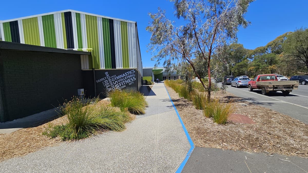 A concrete path from the car park leads to the entrance marked with bright blue line markings. On the left is the venue building, featuring brick walls with an alternating color design. A large banner is affixed to the left wall. Both sides of the pathway are adorned with landscape trees and ornamental plants. On the right is the main road with a few cars in motion.